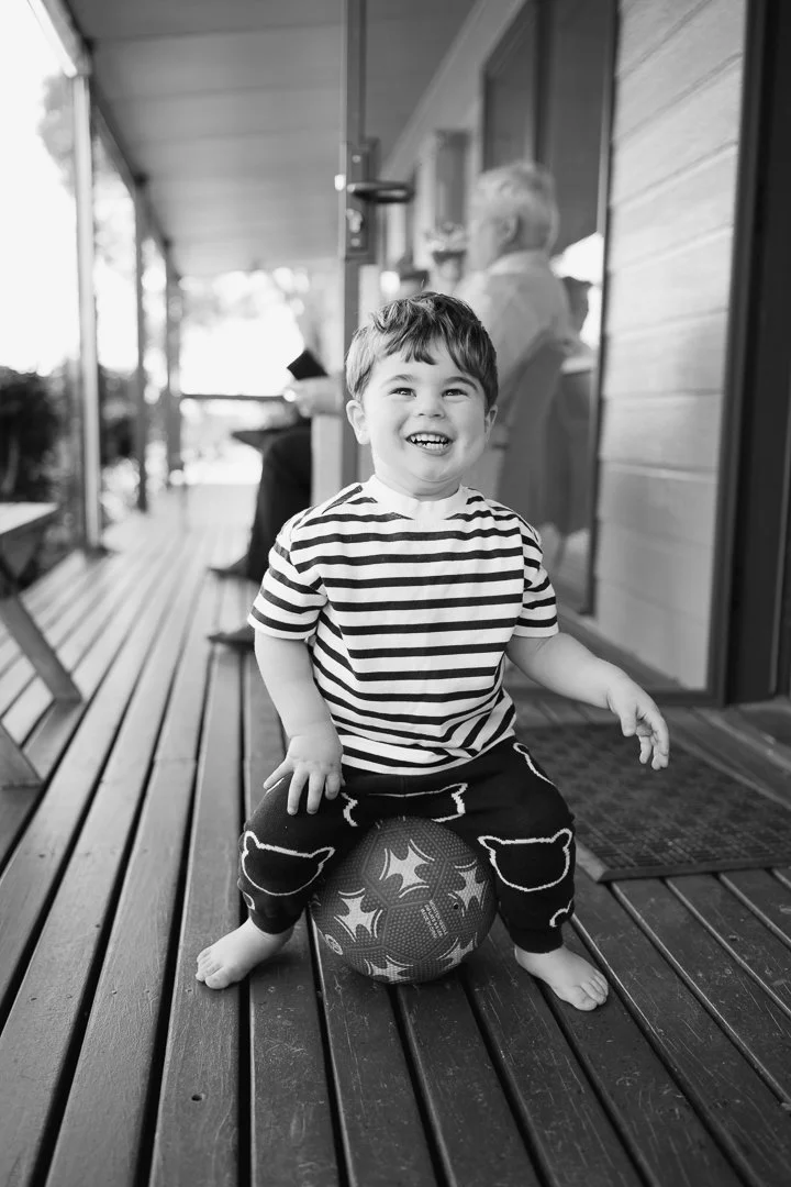 A young boy wearing a striped shirt and patterned pants is sitting on a basketball on a wooden porch, smiling happily at the camera.