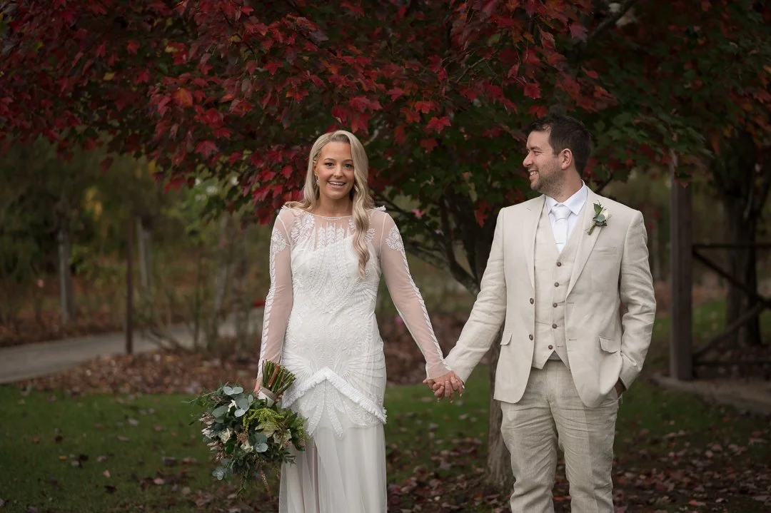 A bride and groom holding hands outdoors with autumn trees in the background.