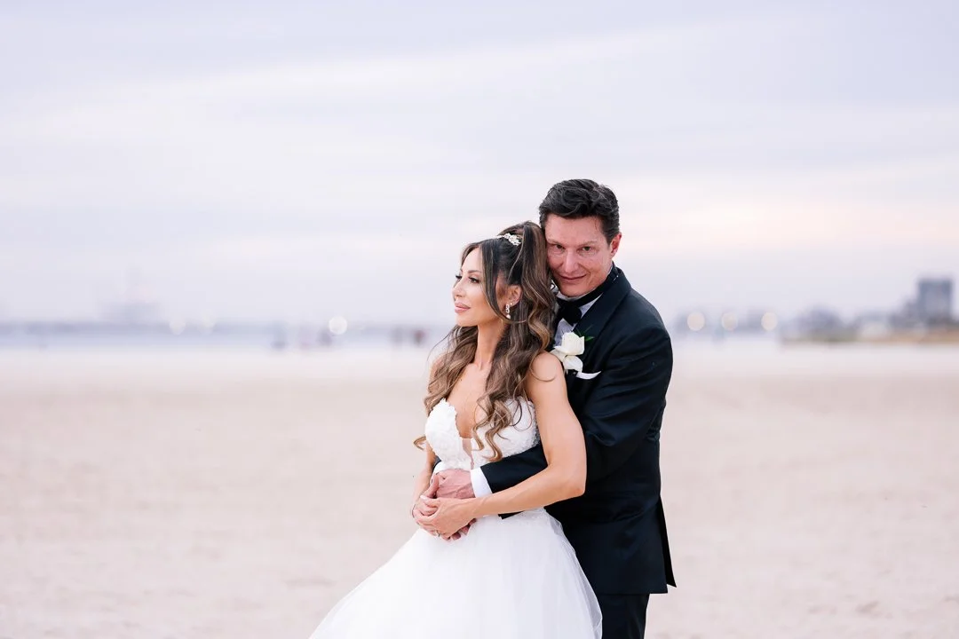 Bride and groom in wedding attire standing on a beach, with the ocean and city buildings in the background.