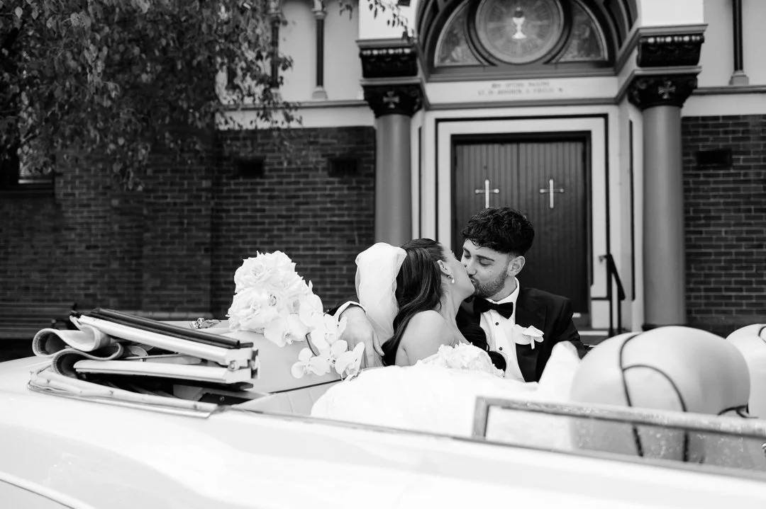 A newlywed couple sharing a kiss in a vintage car in front of a church.