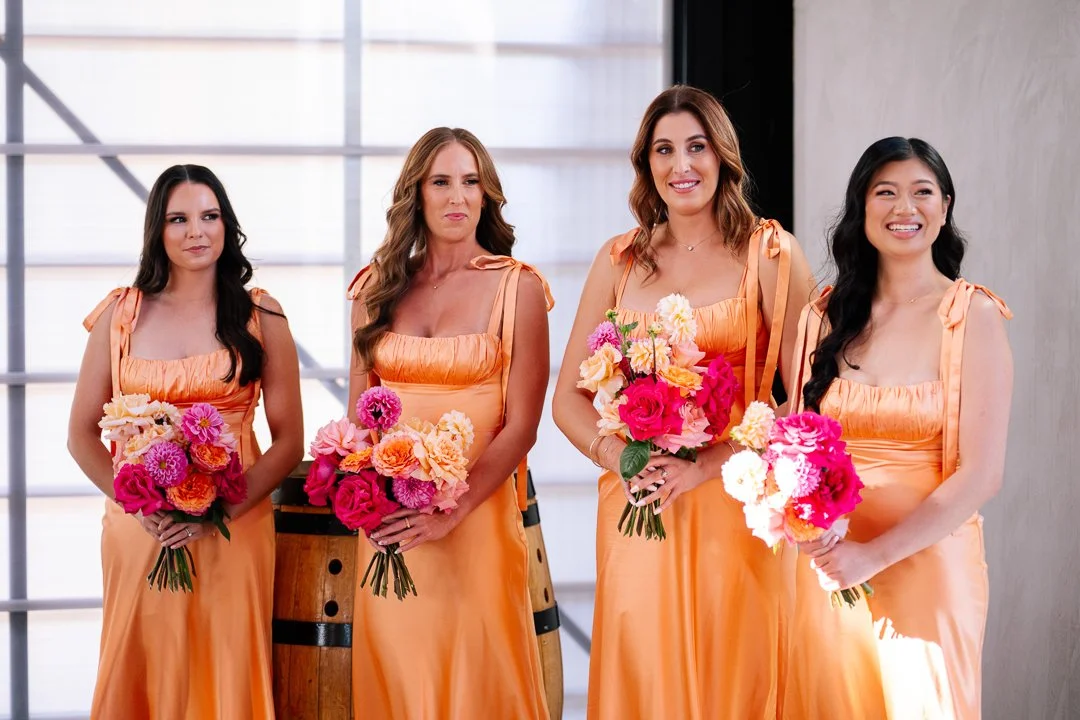 Four women in matching peach-colored dresses holding bouquets of colorful flowers, standing indoors with a large window in the background.