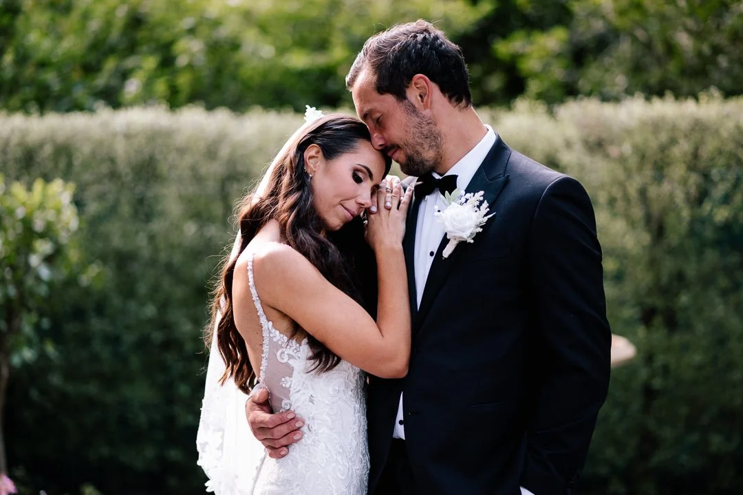 A bride and groom share a romantic moment outdoors, with the bride resting her head on the groom's forehead, and both eyes closed.