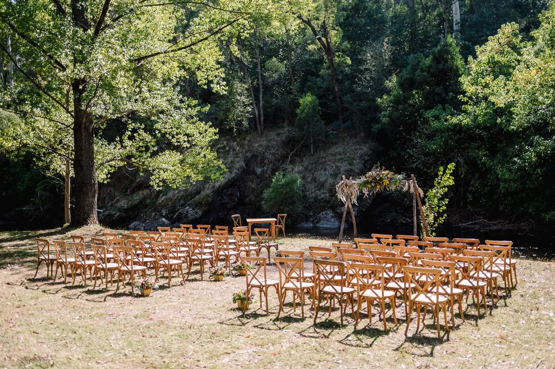 Outdoor wedding setup with rows of wooden chairs arranged in front of a floral arch near a river, surrounded by trees.