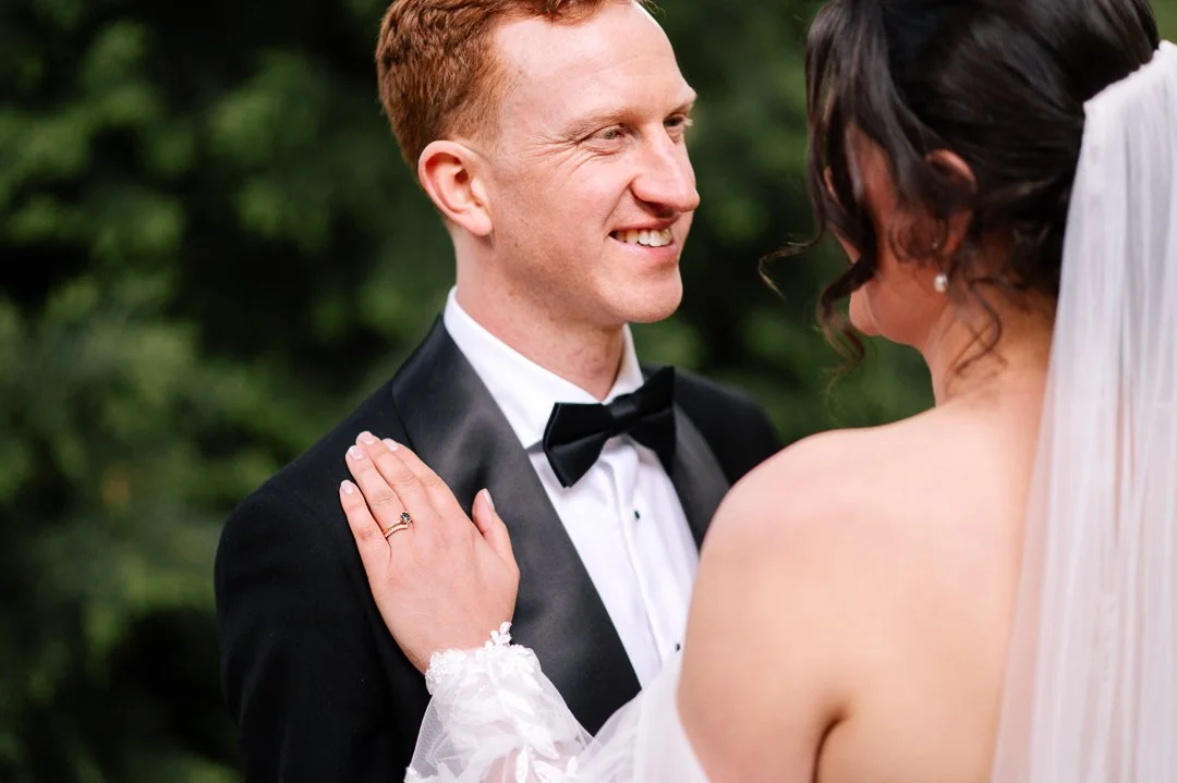 A bride and groom on their wedding day, with the groom smiling and the bride touching his shoulder, outdoors with green foliage in the background.