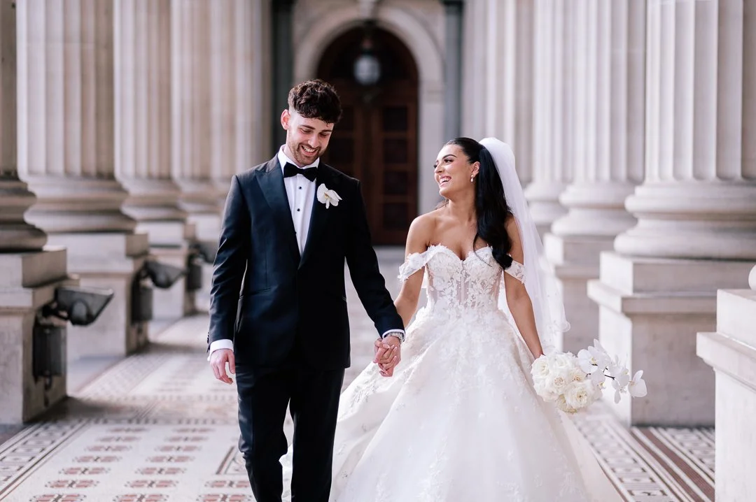 A bride and groom walking hand in hand inside a grand, columned building, smiling happily.