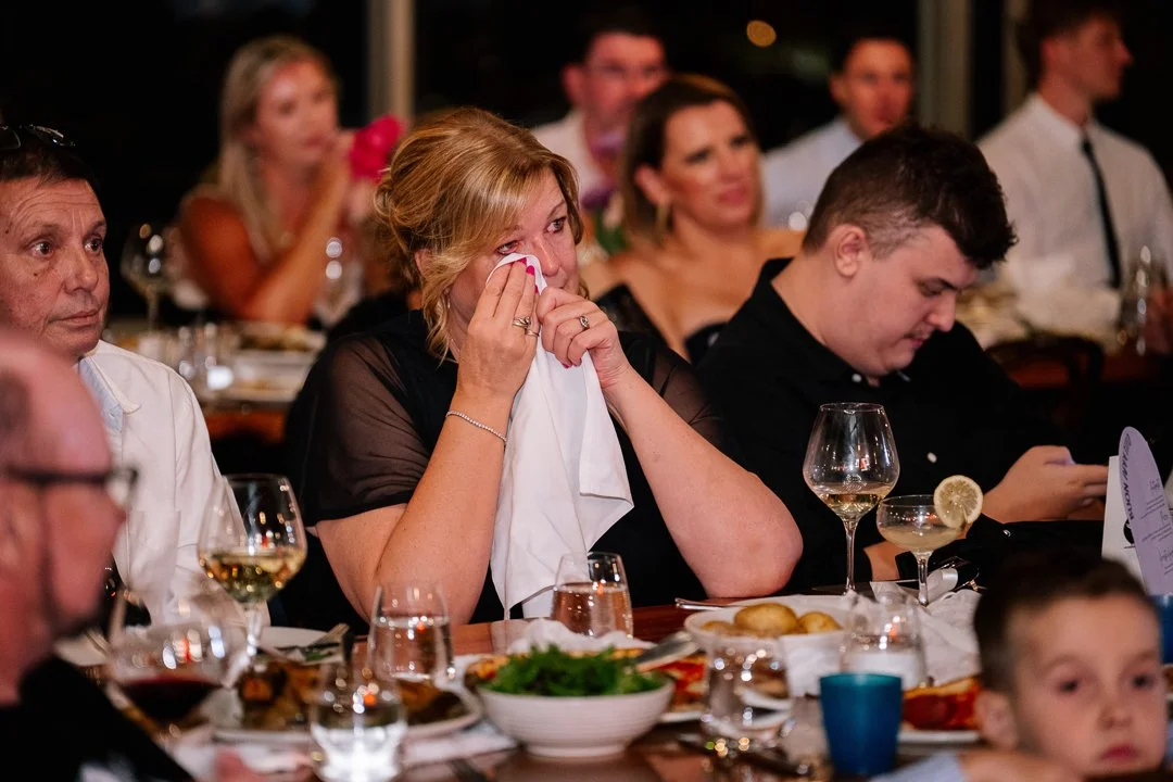 A woman wiping tears from her eyes with a cloth at a formal dinner event, surrounded by other guests at a long table with food and drinks.