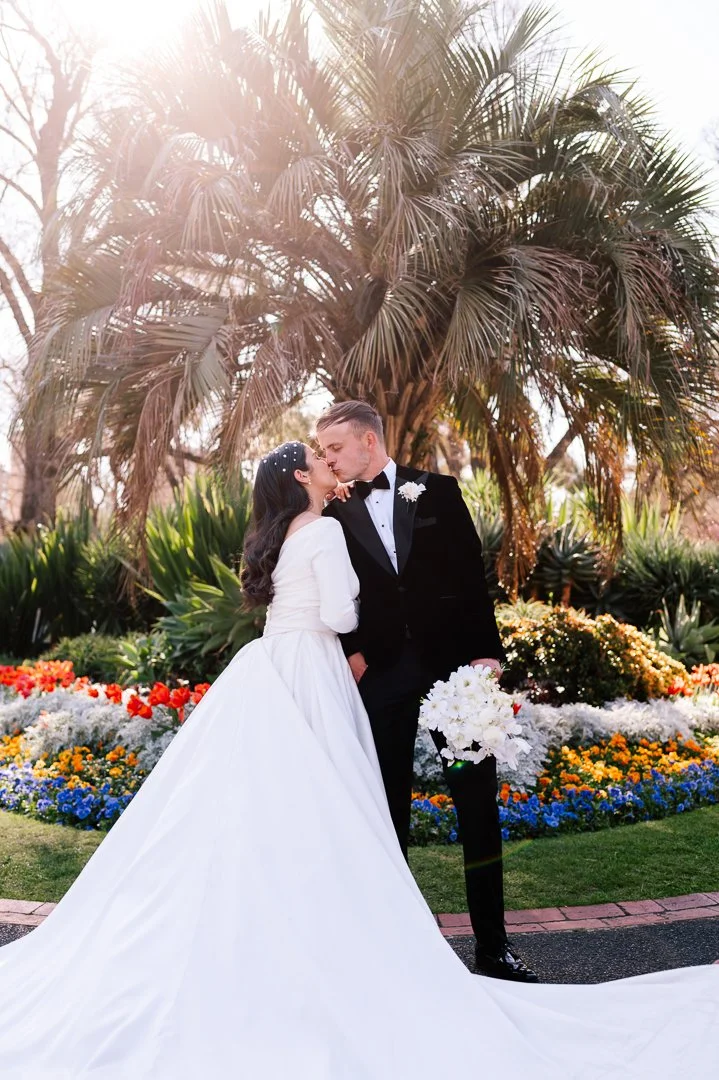 A bride and groom share a romantic moment outdoors during their wedding, with lush greenery and colorful flowers in the background.