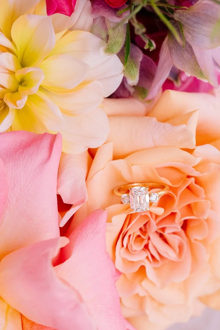 A close-up of a diamond engagement ring placed on a peach rose surrounded by pink and peach roses and soft yellow flowers.
