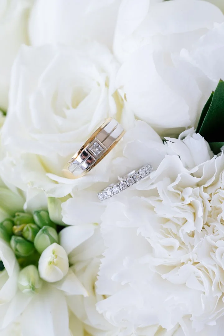 Close-up of wedding rings, one with a single diamond and the other with multiple diamonds, resting on white flowers including roses and hydrangeas.