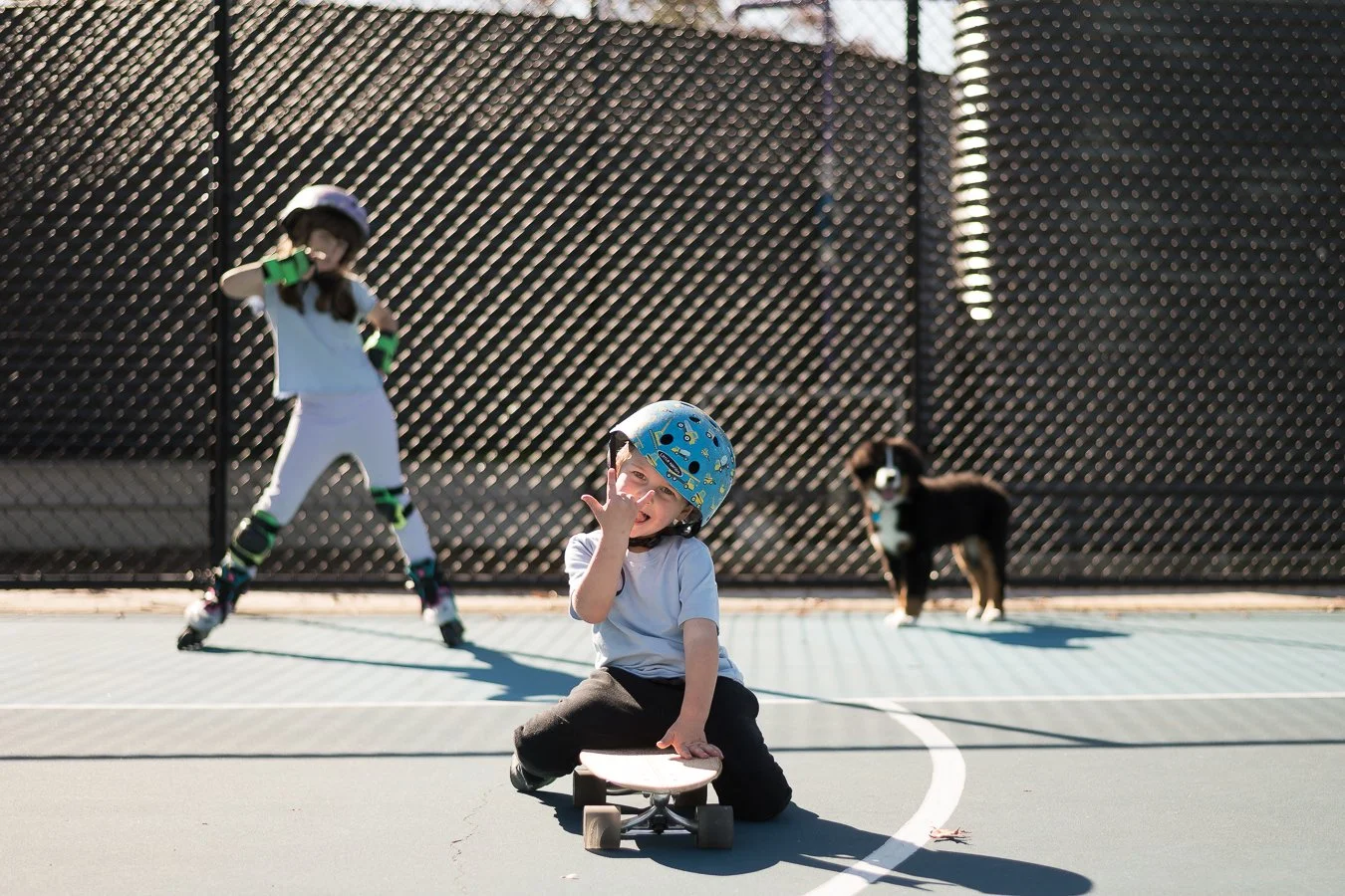 A young boy wearing a helmet and white shirt sitting on a skateboard on a tennis court, with two children rollerblading and a dog in the background near a chain-link fence.