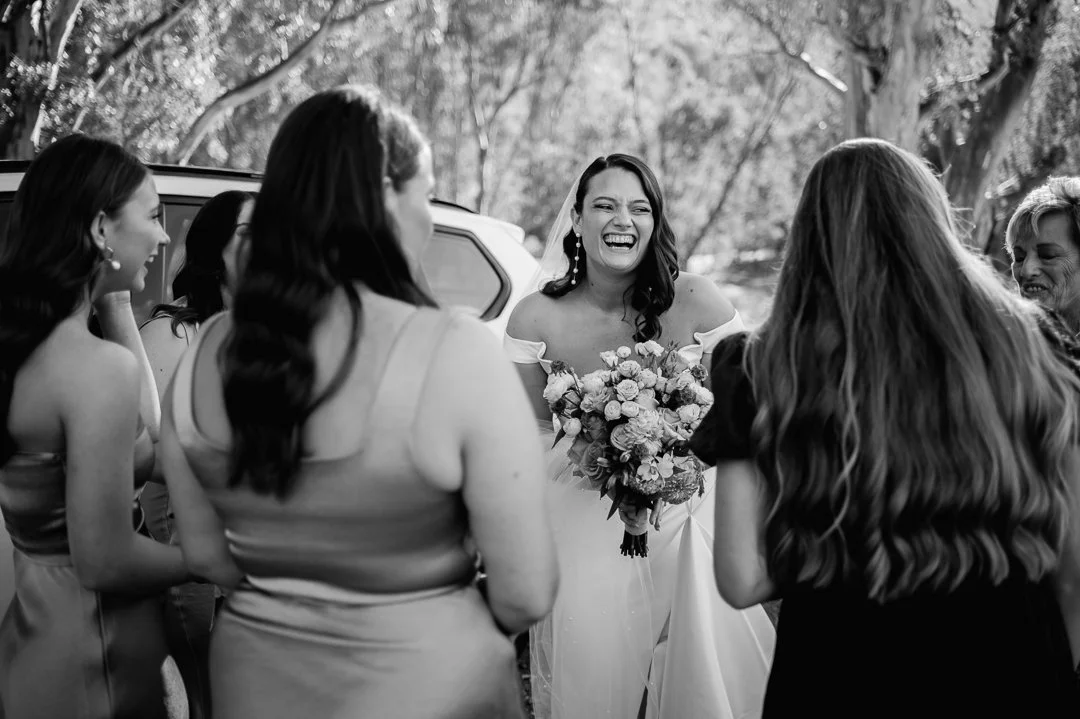 A bride in a wedding dress holding a bouquet of flowers, laughing joyfully with a group of women surrounding her outdoors near trees.