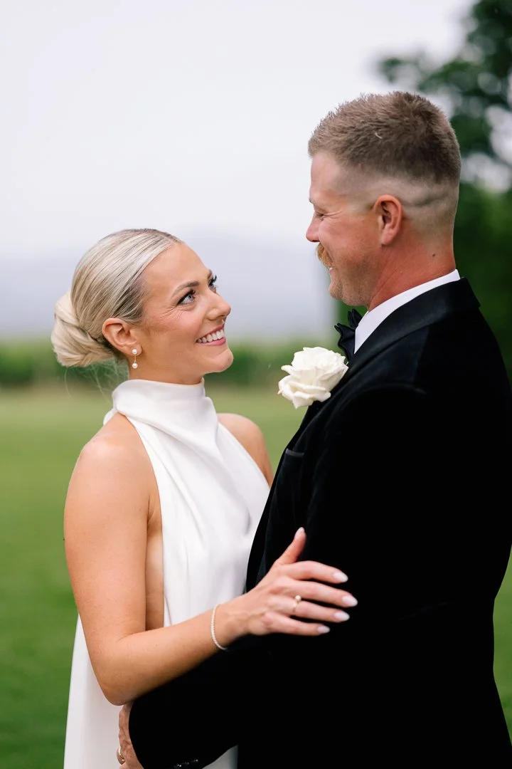 A bride and groom facing each other outdoors, smiling warmly, with the bride in a white dress and the groom in a black tuxedo with a white flower boutonniere.