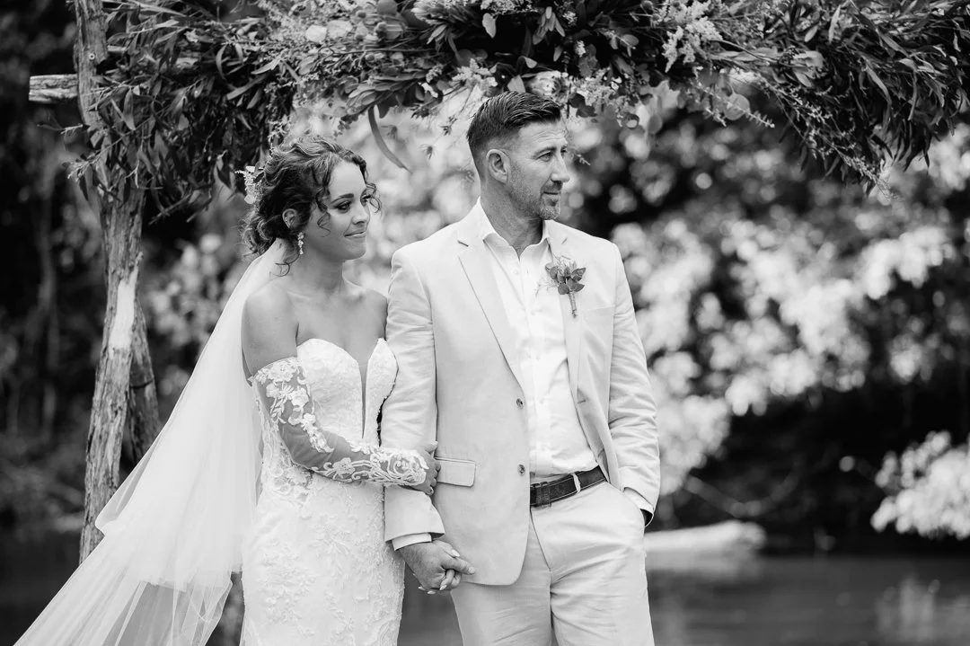 Black and white photo of a bride and groom holding hands under an arch of flowers during their wedding ceremony outdoors.
