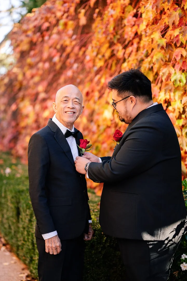 Two men in tuxedos with red roses in their lapels standing outdoors in front of an orange and red autumn leaf-covered wall. One man is helping the other with his rose, and both are smiling.