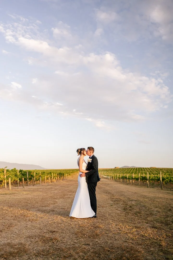 A bride and groom in wedding attire embracing in a vineyard at sunset.