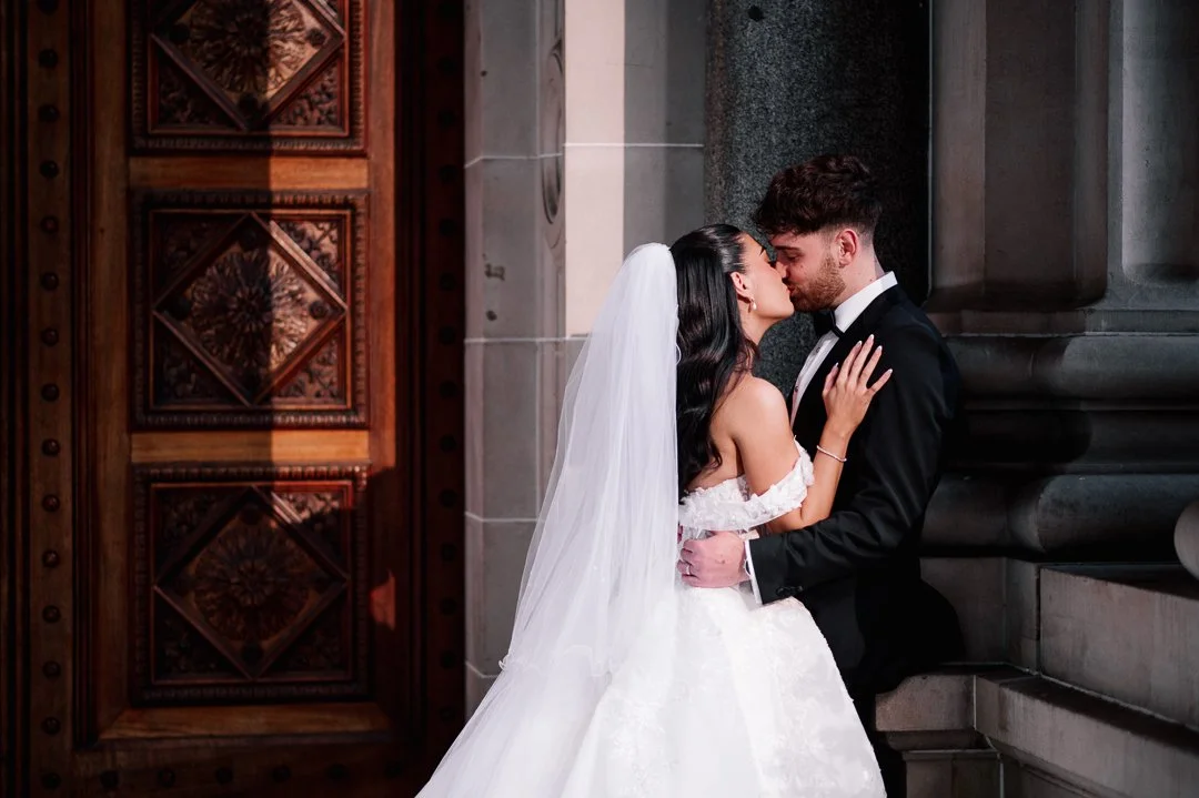 A bride and groom sharing an intimate moment, about to kiss, inside a grand stone building with elaborate wooden door.