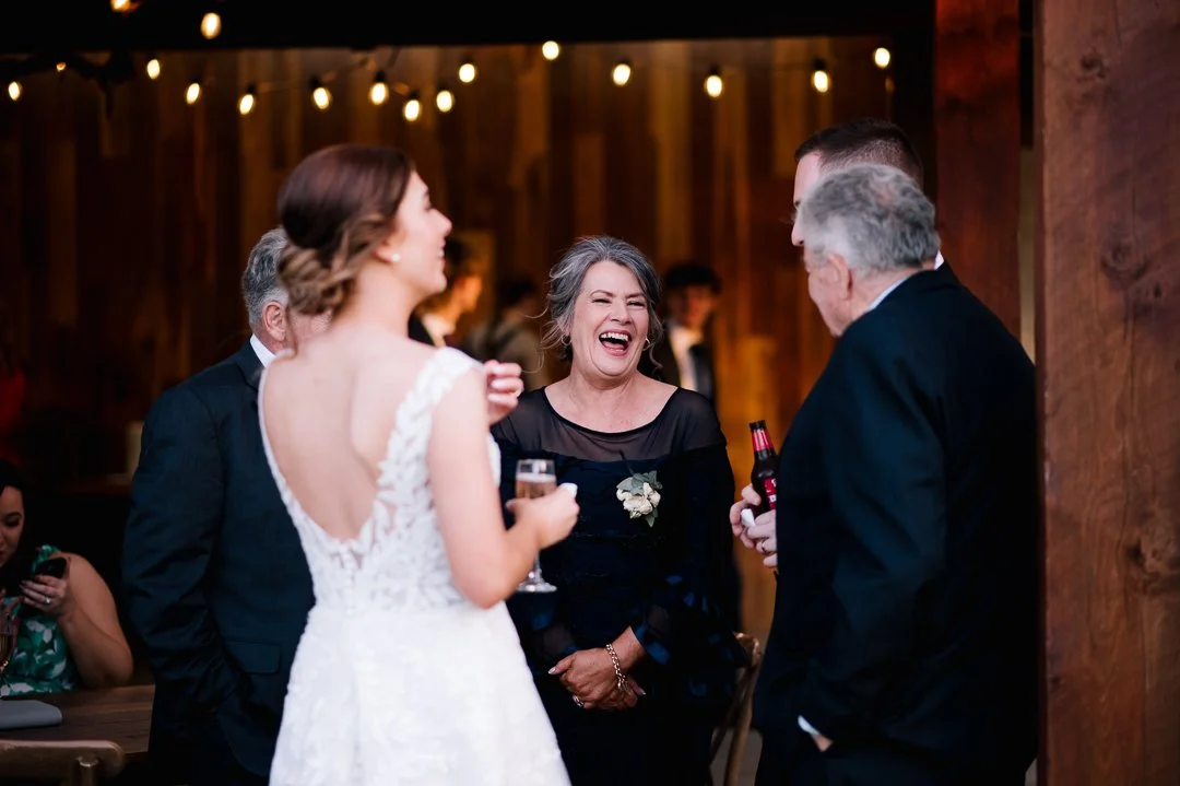 Wedding reception with a bride in a white dress talking to a woman in a black dress, all smiling and holding drinks. Two men in suits are also present, one holding a beer and the other with a hand in his pocket. Warm lighting and wooden decor create