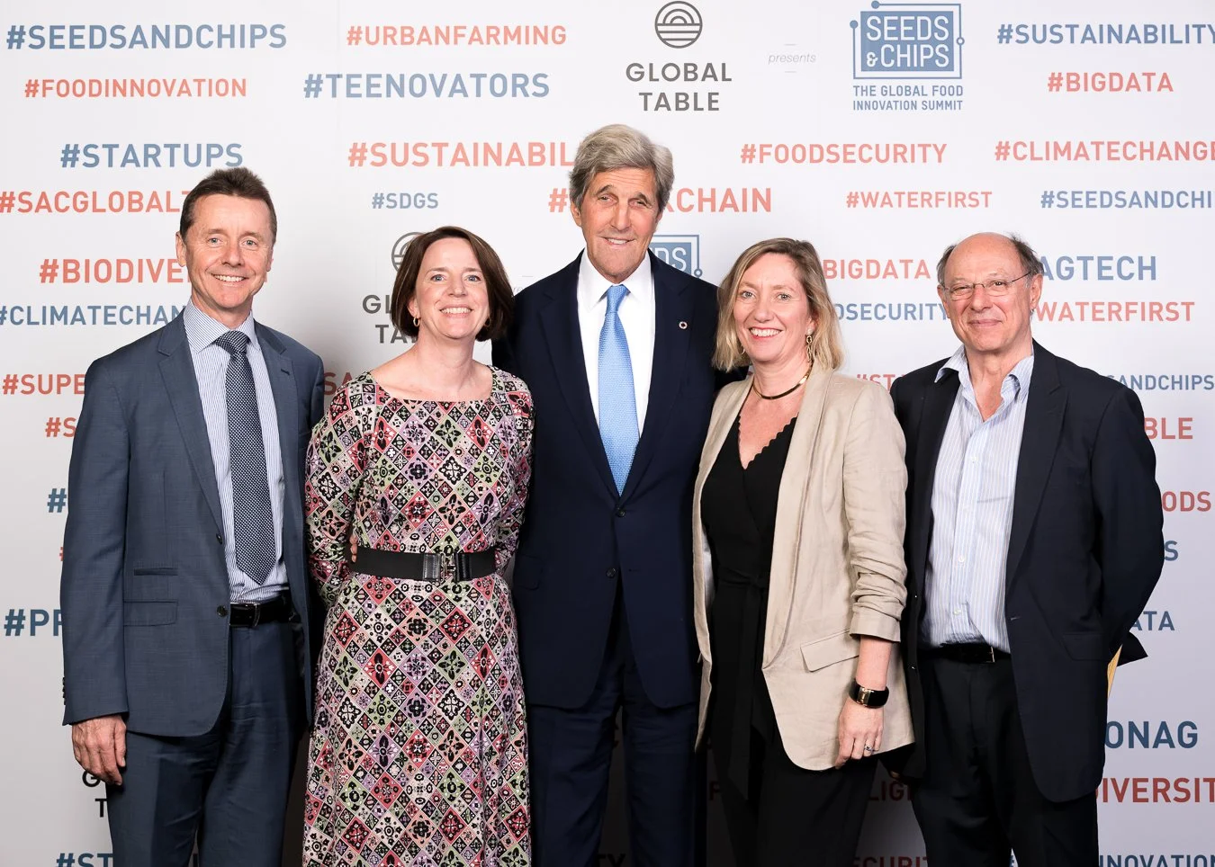 Group of five professionally dressed people smiling at an event with a backdrop featuring hashtags related to food innovation, sustainability, and global food topics.