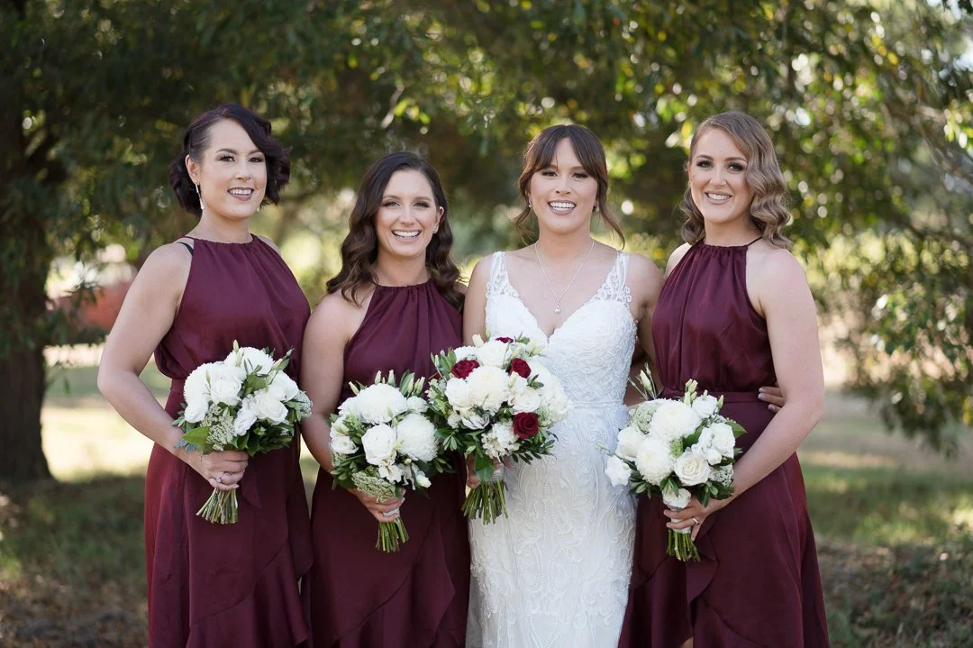 Bride in white wedding dress standing with three bridesmaids in burgundy dresses, outdoors with trees in the background, each holding a bouquet of white and red flowers.