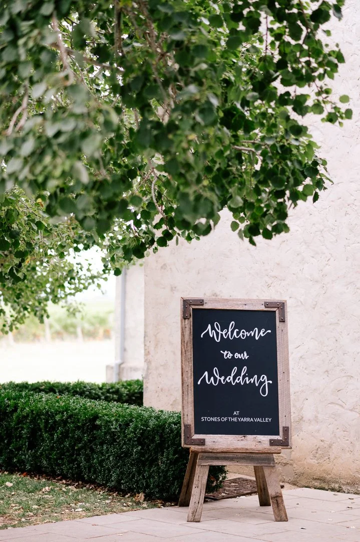 A wooden framed chalkboard sign on an easel reading "Welcome to our Wedding at Stones of the Yarra Valley," placed outdoors near greenery and a white stucco wall.