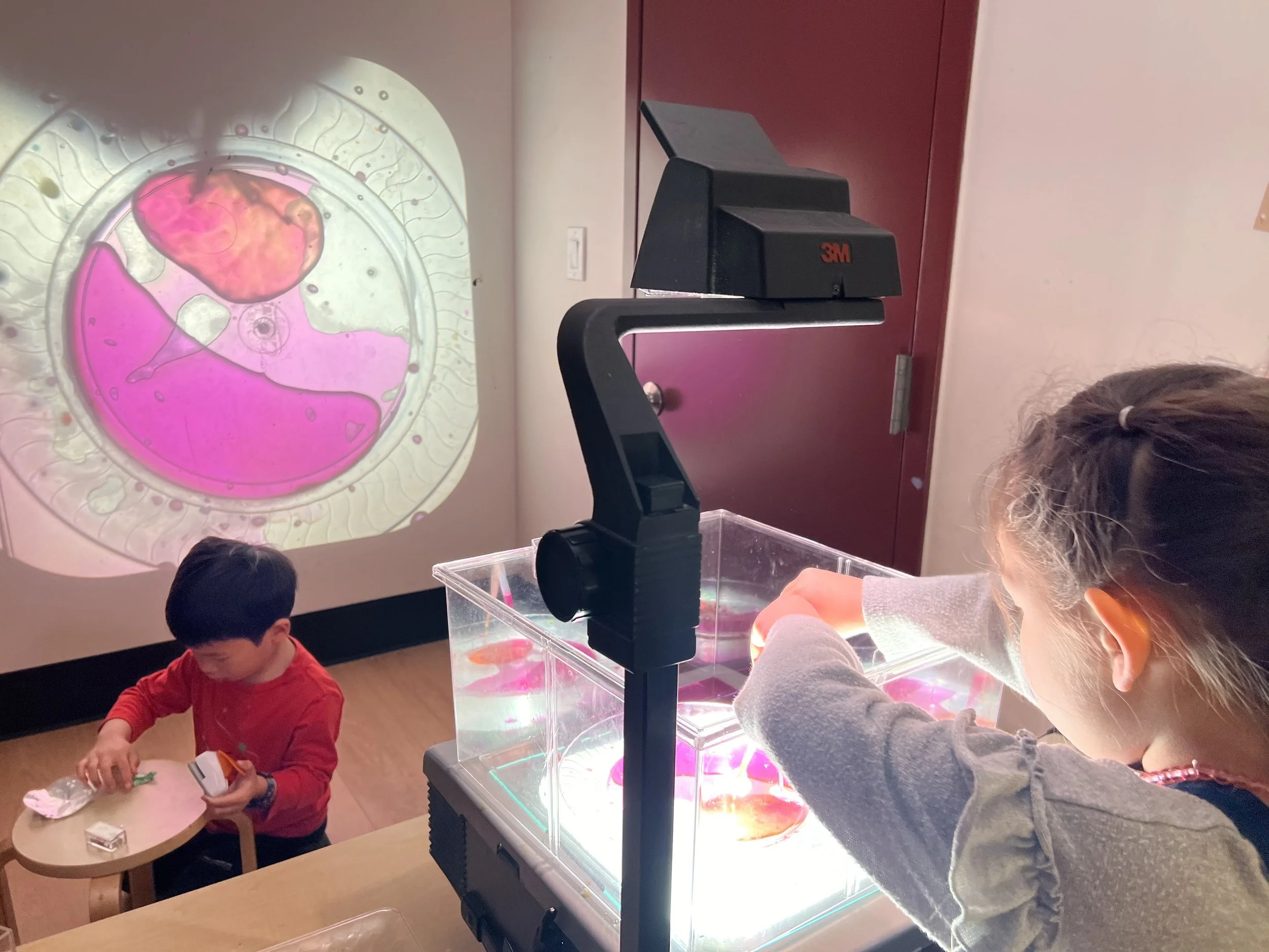 Two children observing a petri dish with live aquatic creatures, using a microscope at an indoor educational exhibit.