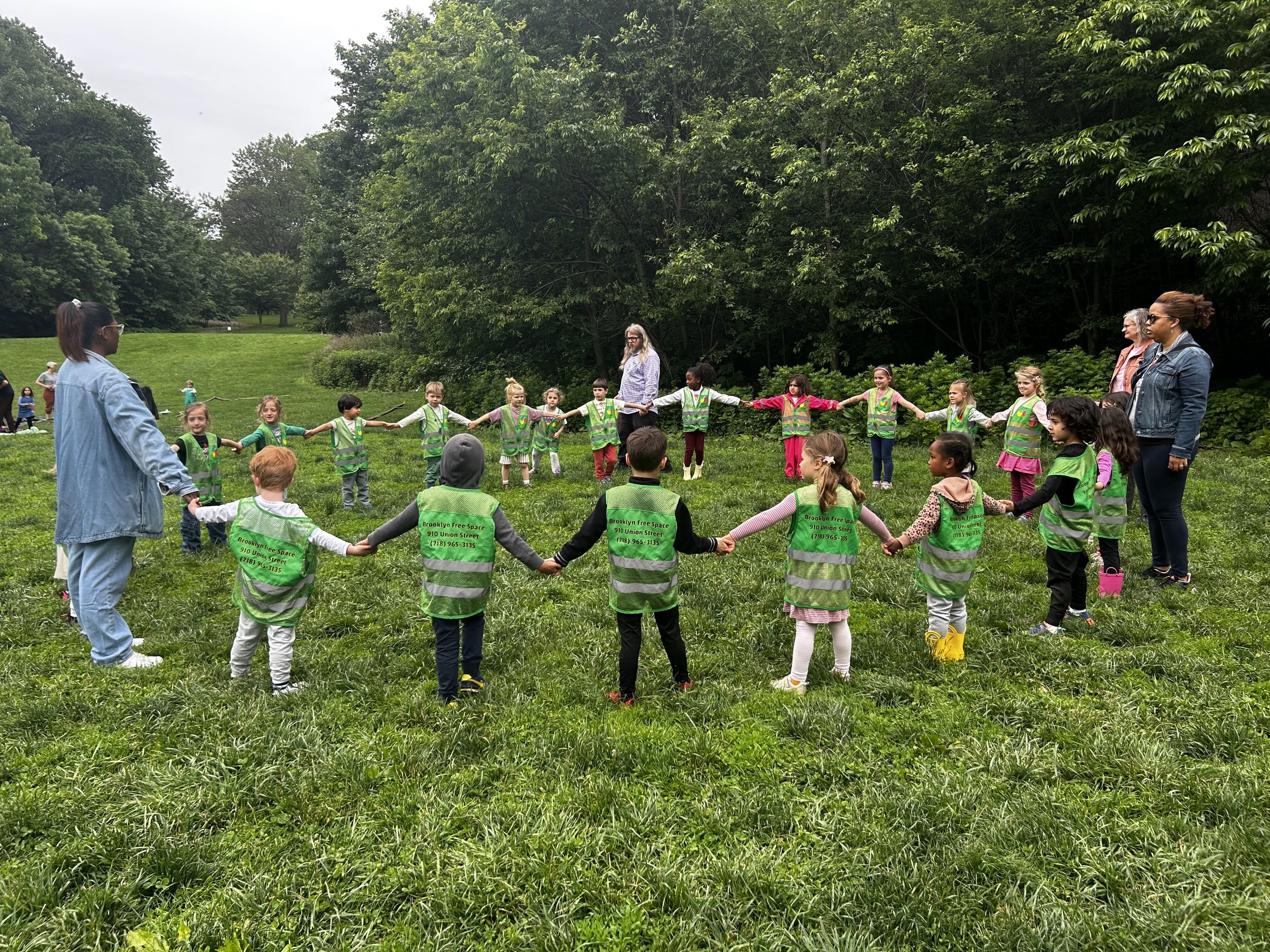 Children and adults form a large circle holding hands in a grassy park, with trees in the background during an outdoor activity.