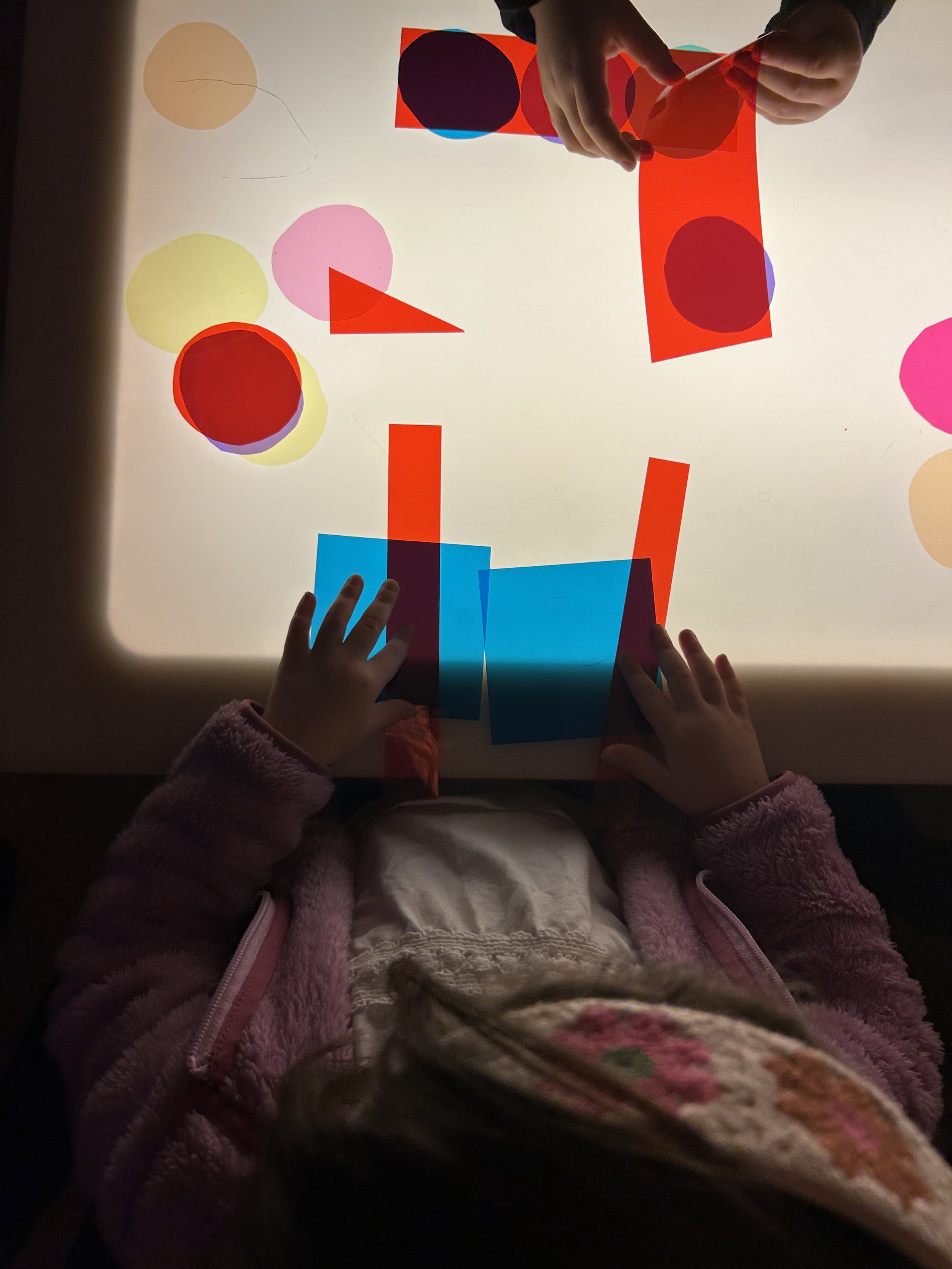 Children's hands interacting with a light-up surface displaying colorful geometric shapes including circles, squares, and triangles.