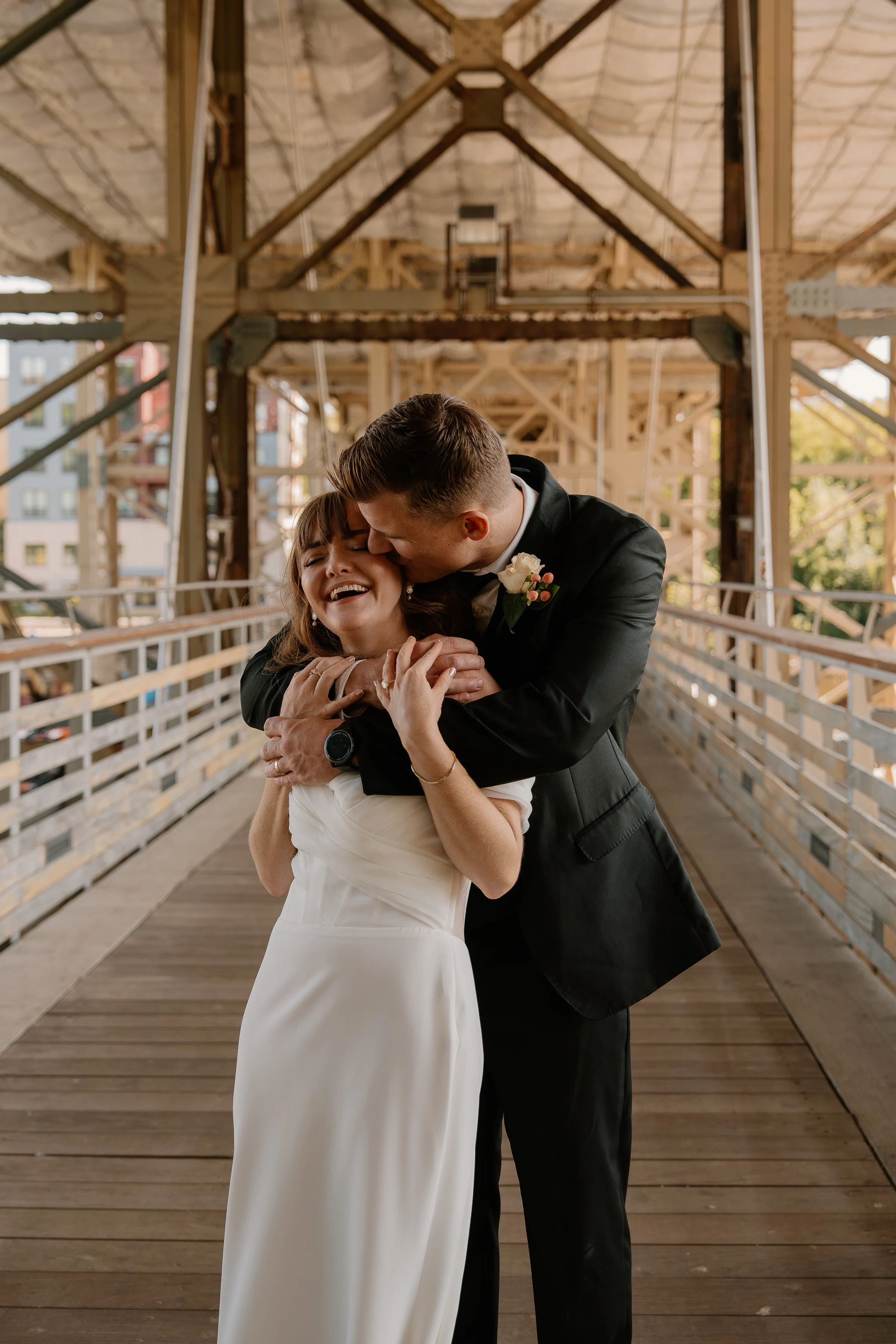 A bride and groom sharing a joyful embrace on a wooden bridge, with the groom kissing the bride's head, during their wedding.