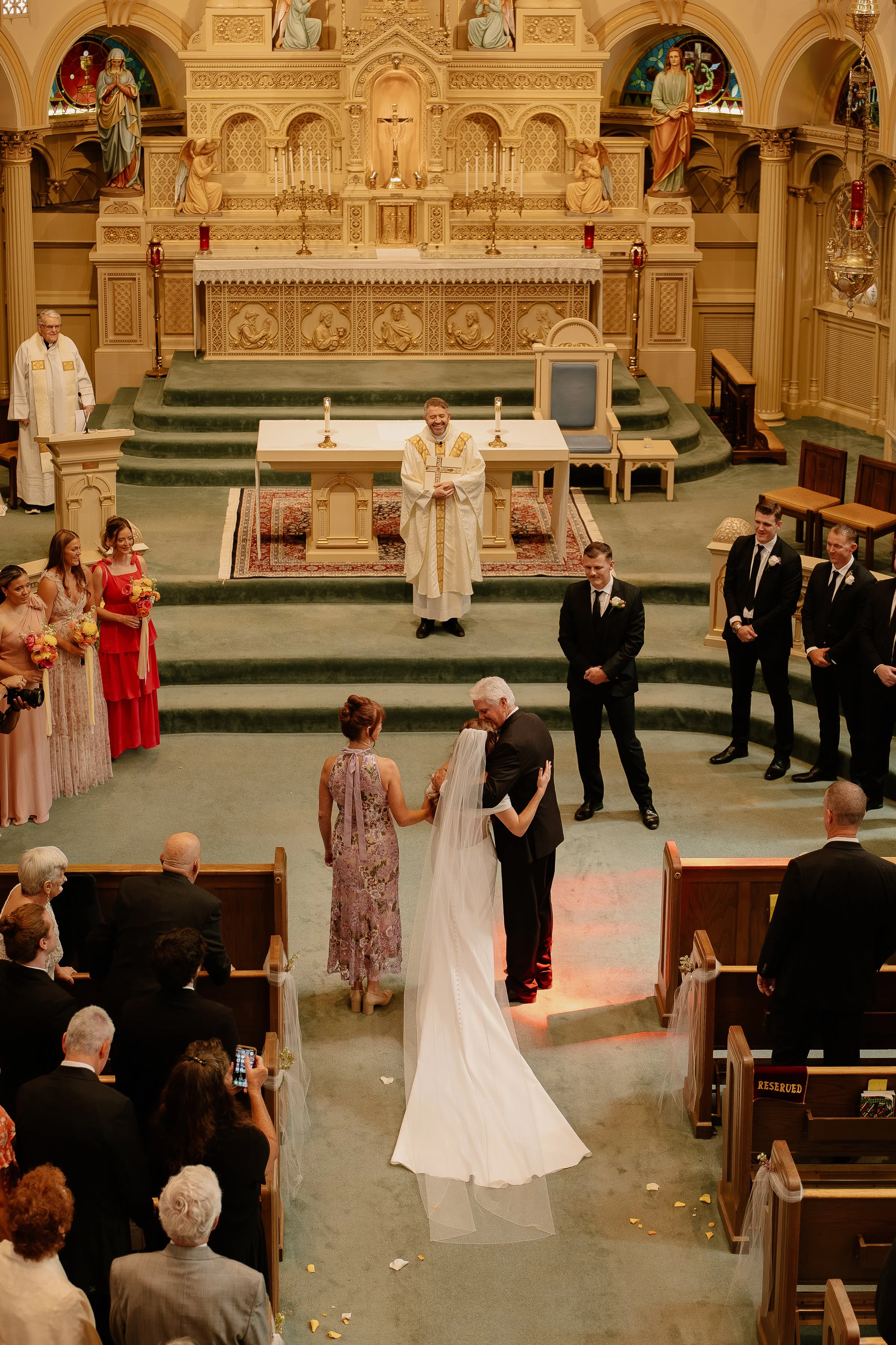A wedding ceremony taking place inside a church, with a priest standing behind the altar, a bride and groom hugging at the front, surrounded by bridesmaids in pastel dresses and groomsmen in black suits, and guests seated in pews.