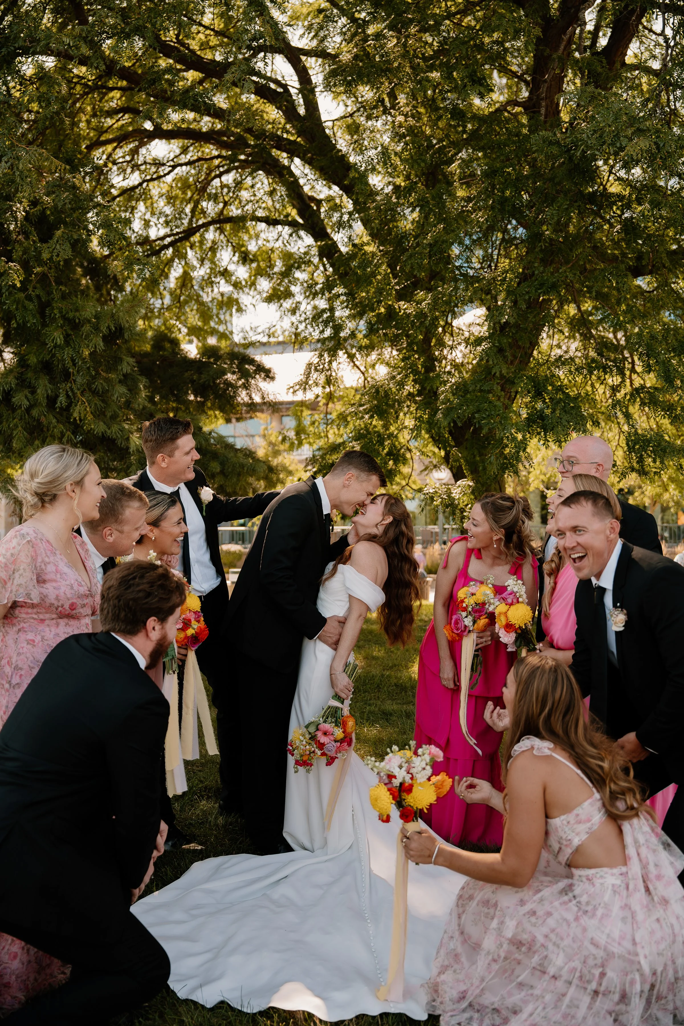 A group of wedding guests surrounds a bride and groom outdoors, celebrating under a large tree, with cheerful expressions, bouquets, and a sunny background.