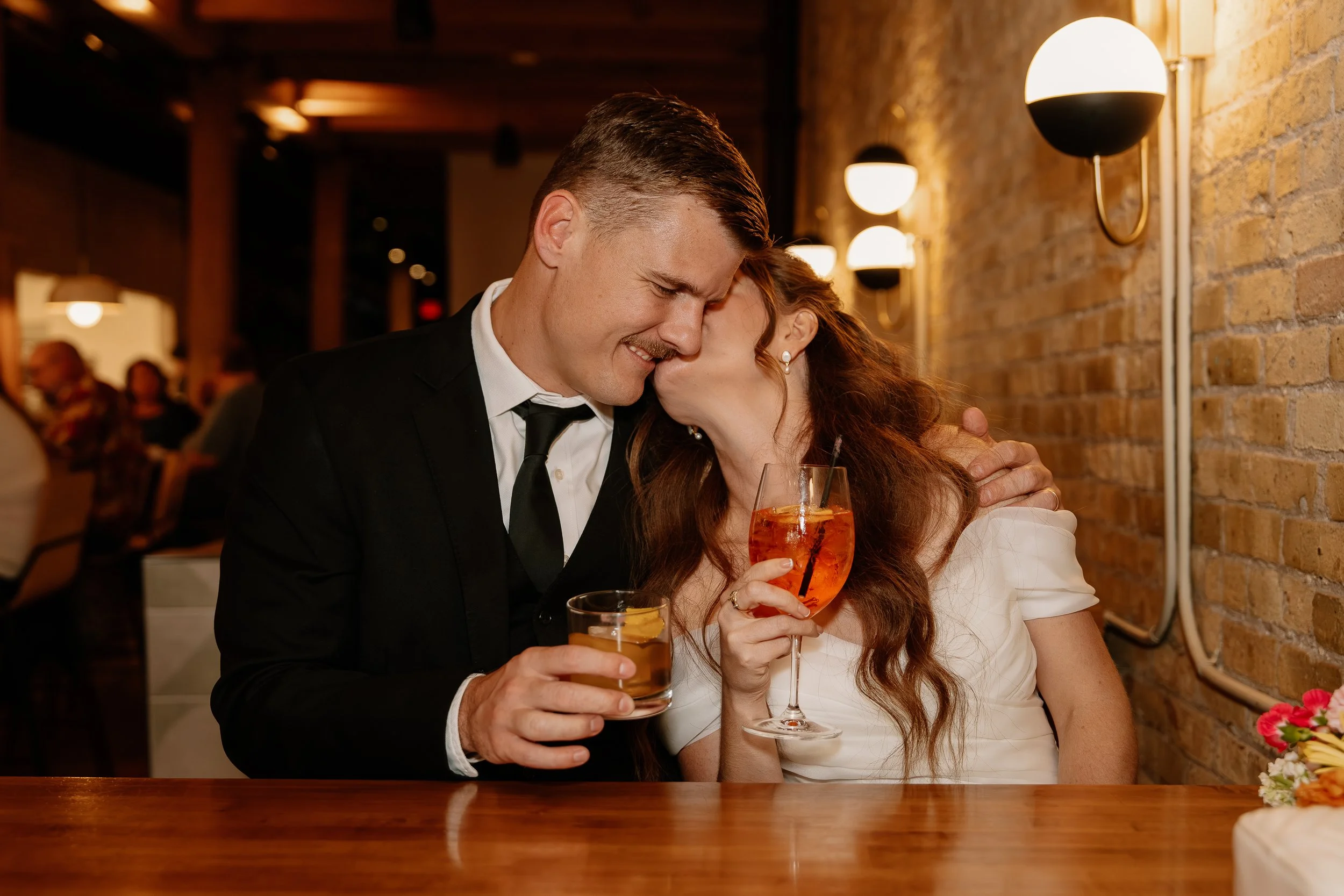 A man and a woman sharing a kiss while holding drinks at a restaurant or bar with brick walls and warm lighting.