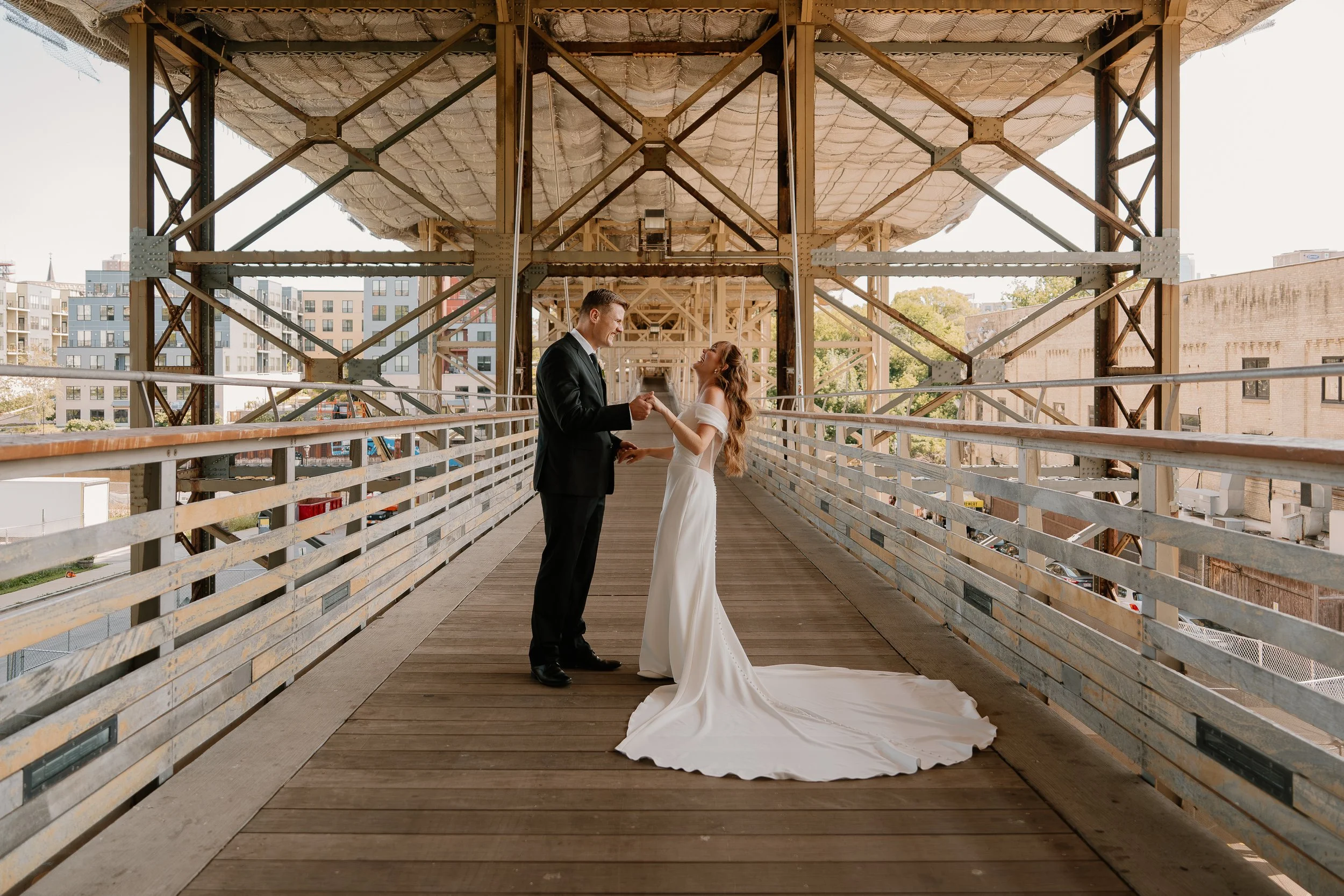 A bride and groom exchanging vows on a wooden bridge in an urban setting, with the groom in a black suit and the bride in a white off-shoulder gown.