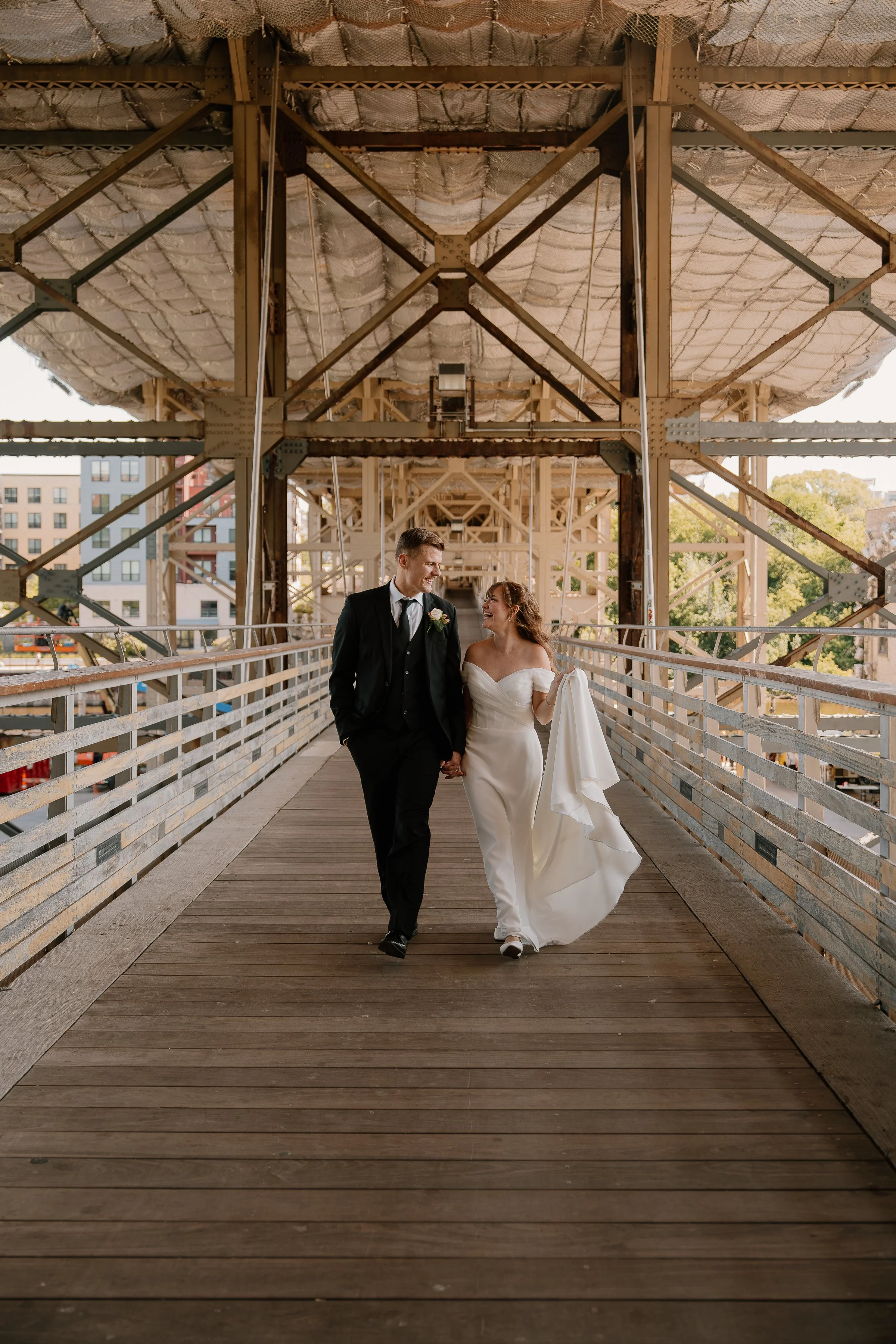 A newlywed couple walking hand-in-hand on a wooden bridge with an industrial structure overhead, with city buildings and trees in the background.