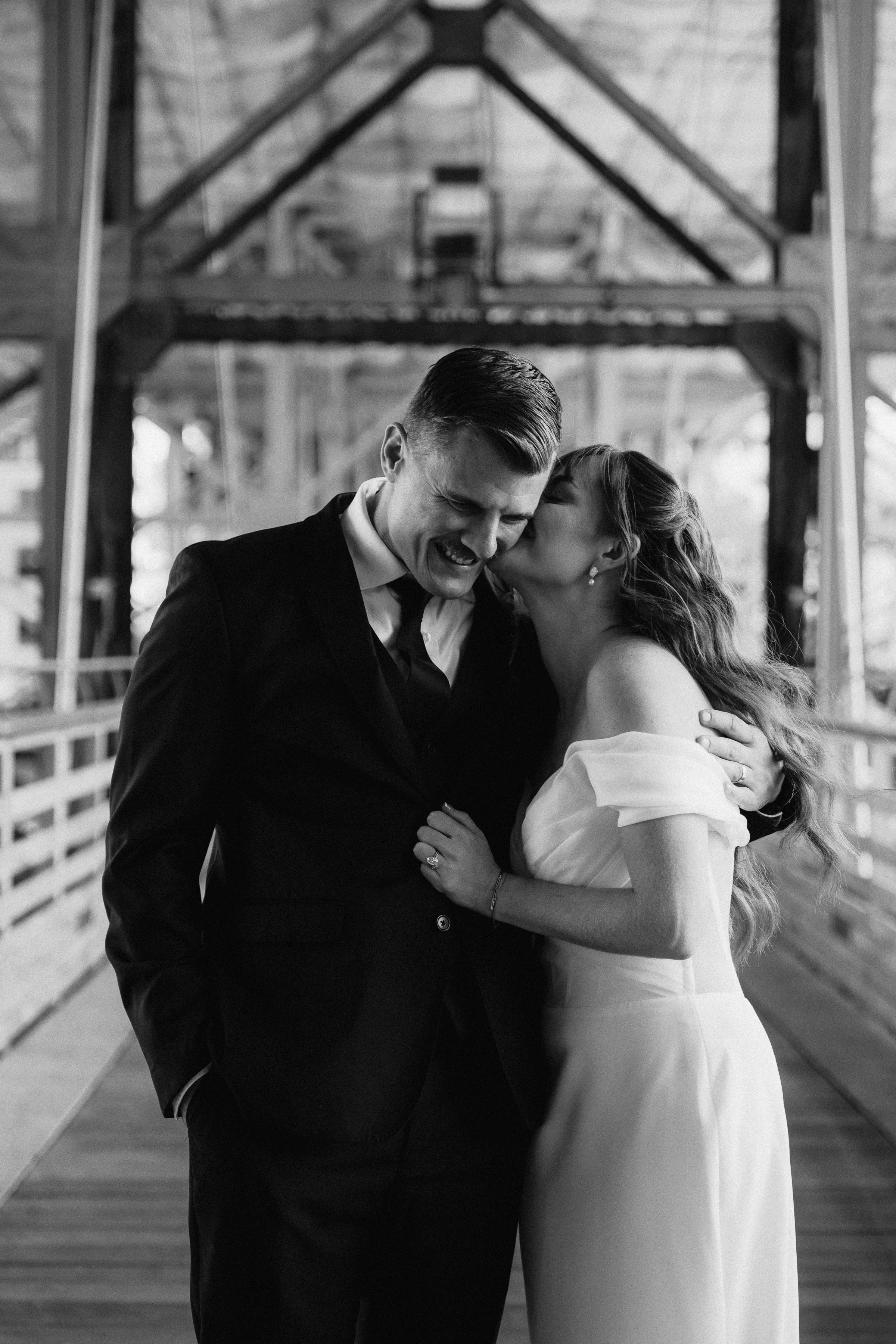 Black and white photo of a bride and groom sharing a joyful moment, with the bride kissing the groom on the cheek inside a rustic wooden barn.