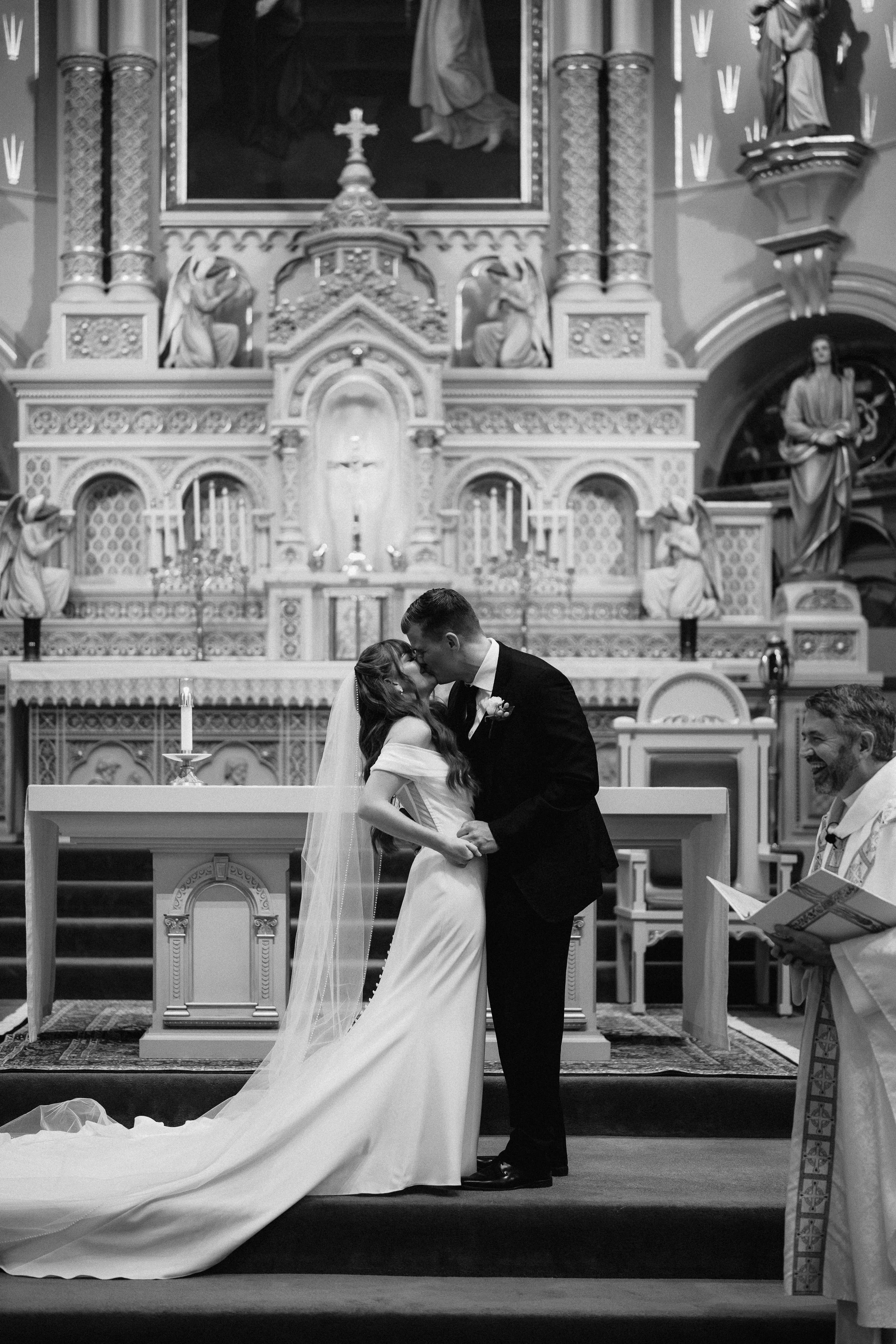 A black and white photo of a wedding ceremony inside a church, showing a bride and groom kissing at the altar, with a priest on the right holding a book, and ornate religious decorations and statues in the background.