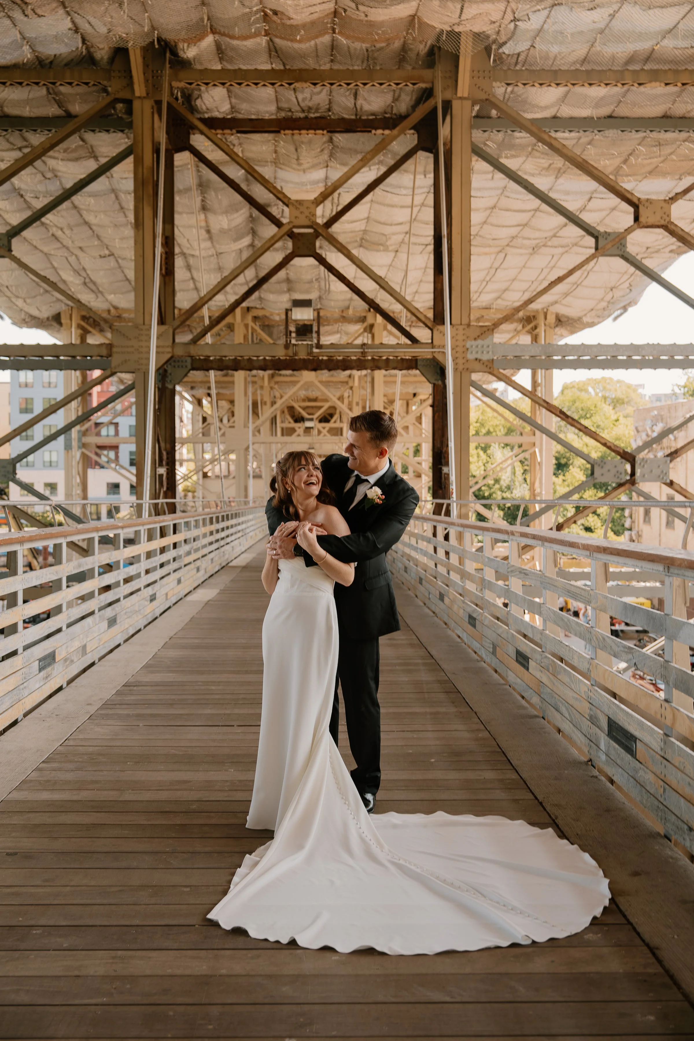 Couple in wedding attire dancing on a bridge with a metal framework and wooden floor, with city buildings and trees in the background.