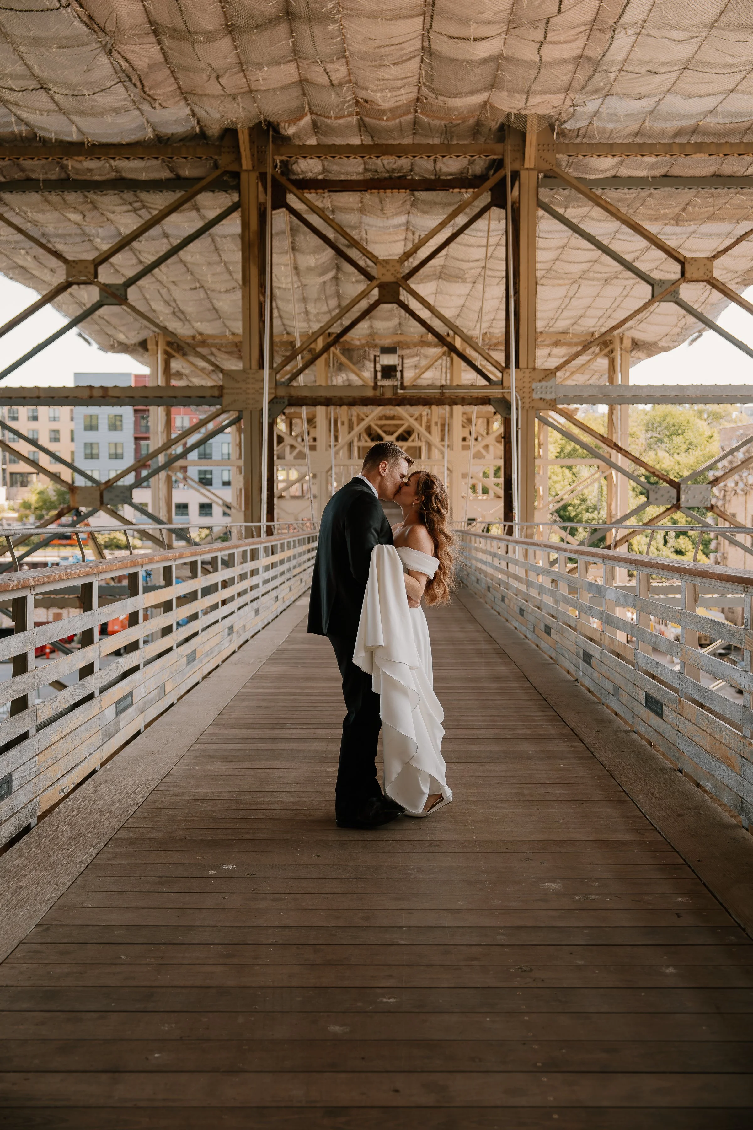 A bride and groom sharing a kiss on a wooden bridge under an industrial structure, with city buildings and trees in the background.