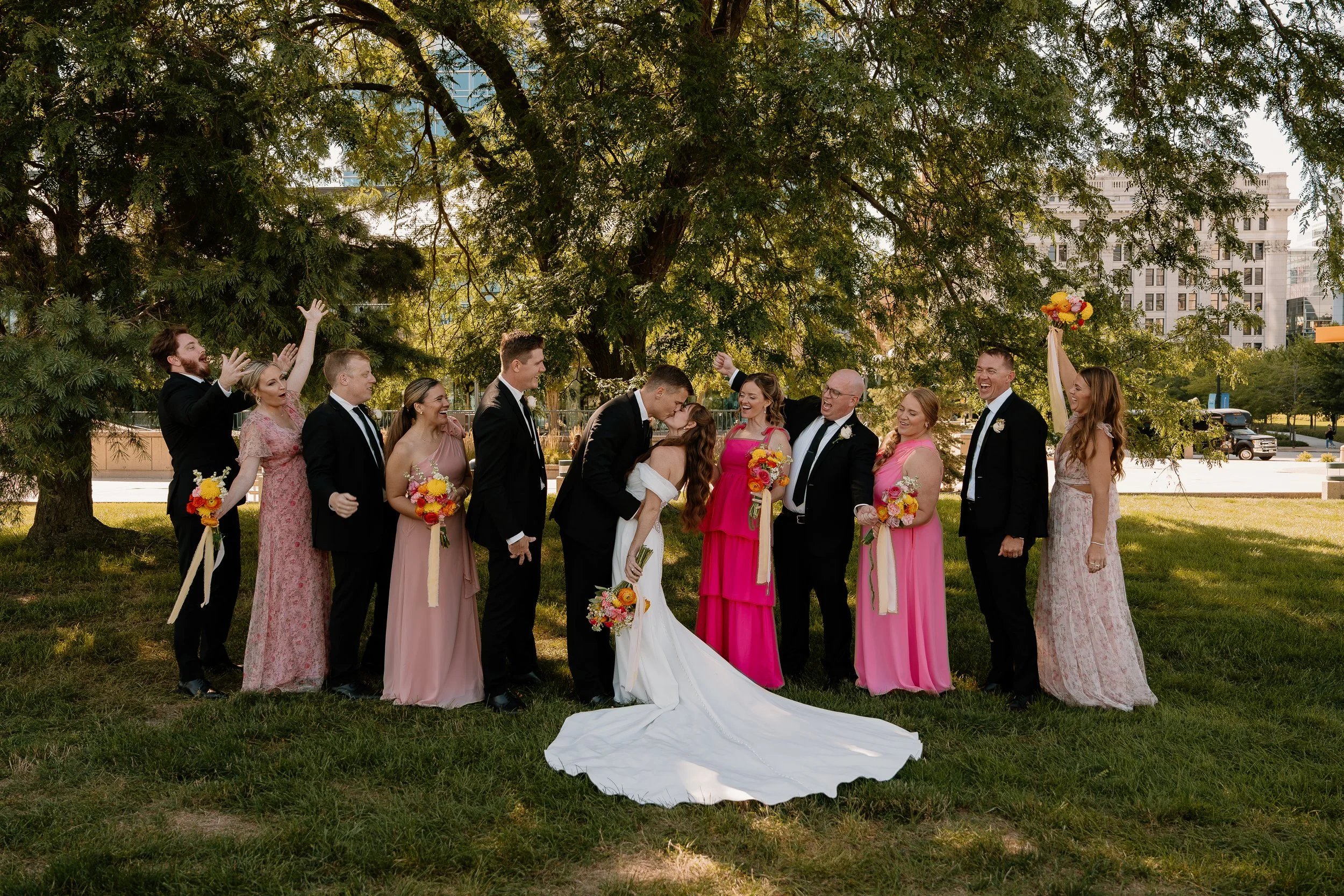A wedding party of twelve people, including the bride and groom, standing outdoors under a large tree. The bride, in a white wedding dress, and the groom, in a black suit, are sharing a kiss in the center. The bridesmaids are dressed in pink gowns, a