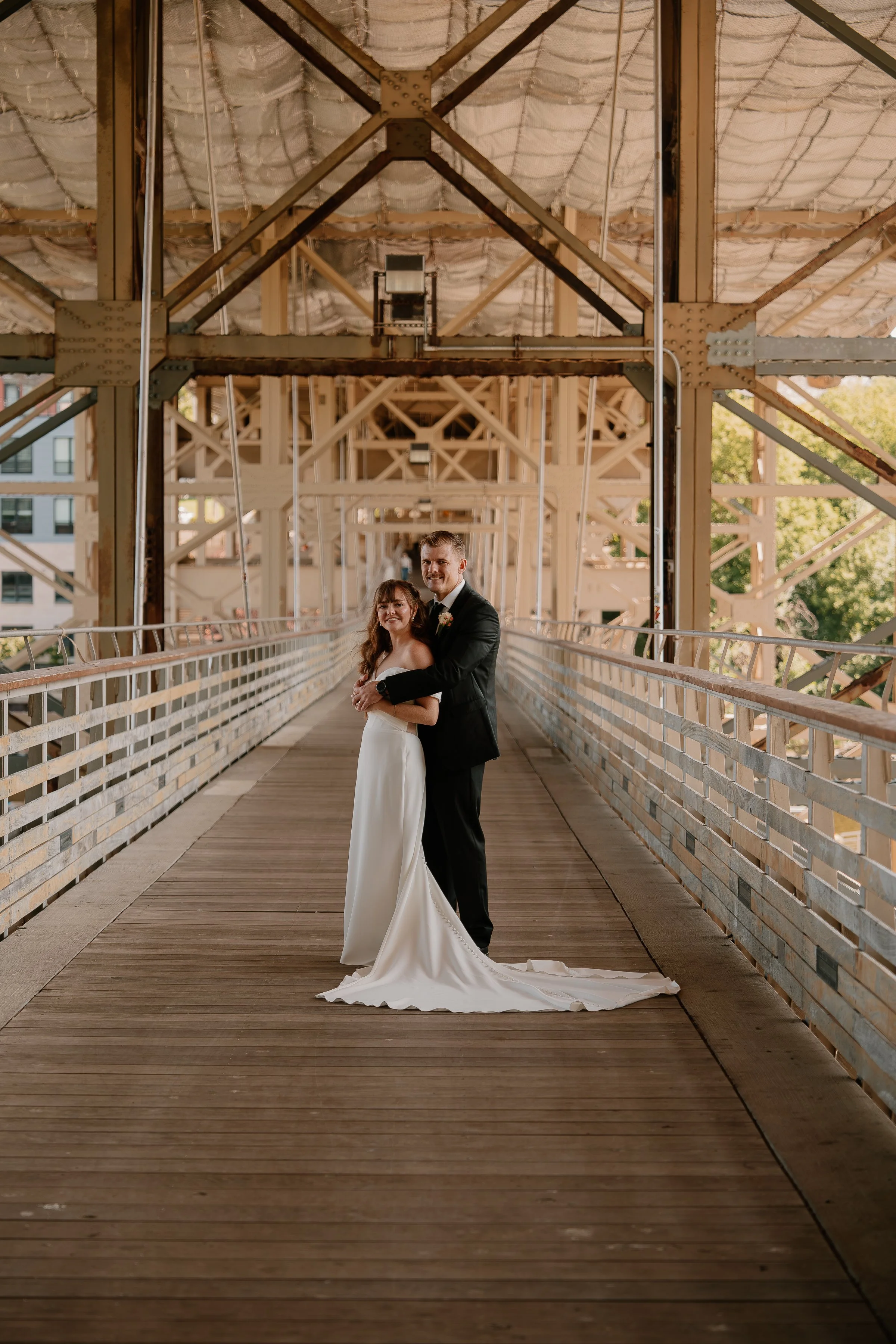A newlywed couple standing on a wooden bridge with industrial and urban background, the bride in a white satin wedding gown and the groom in a black tuxedo, smiling and embracing.