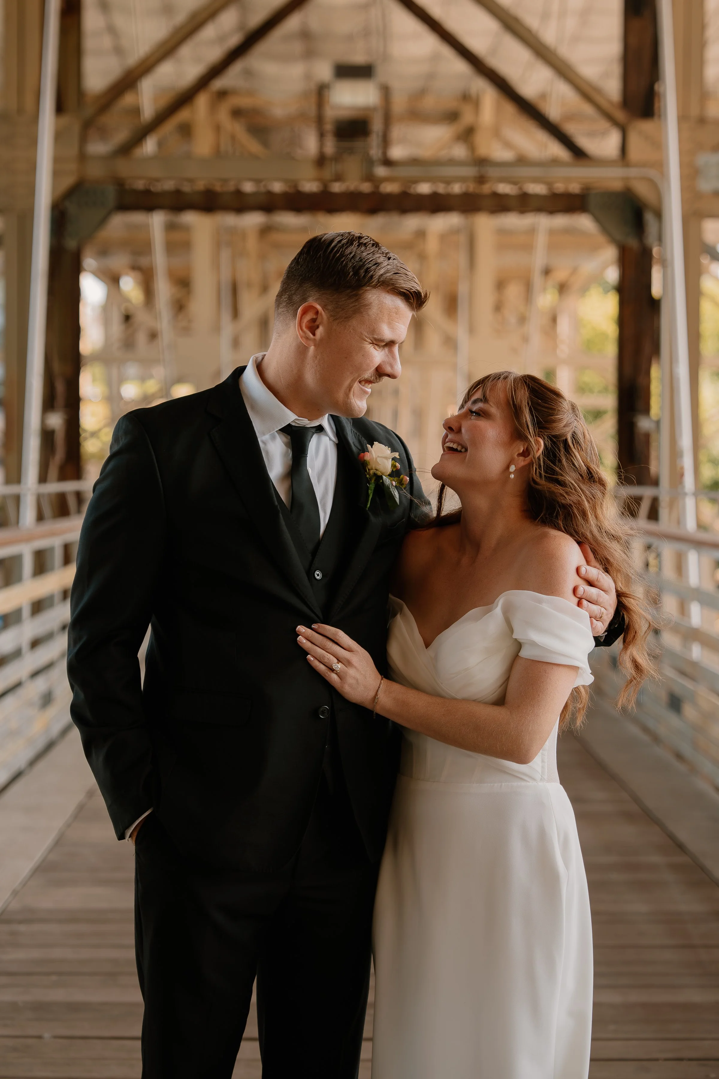 A newlywed couple standing on a wooden bridge inside a rustic barn, smiling and gazing into each other's eyes.