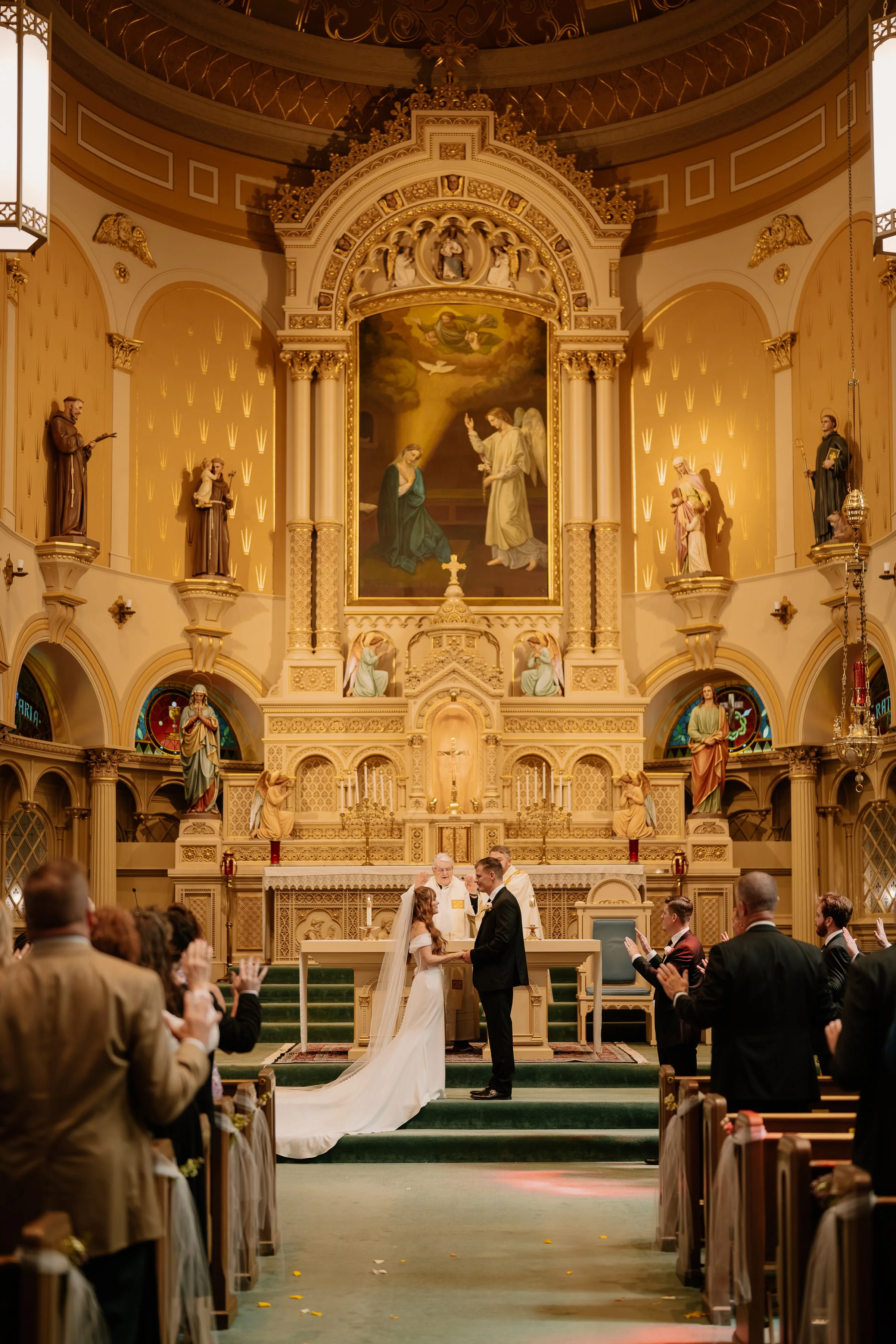 A wedding ceremony taking place inside a church with a large ornate altar and religious artwork in the background. The bride and groom are standing together at the altar, holding hands, surrounded by guests clapping and smiling.