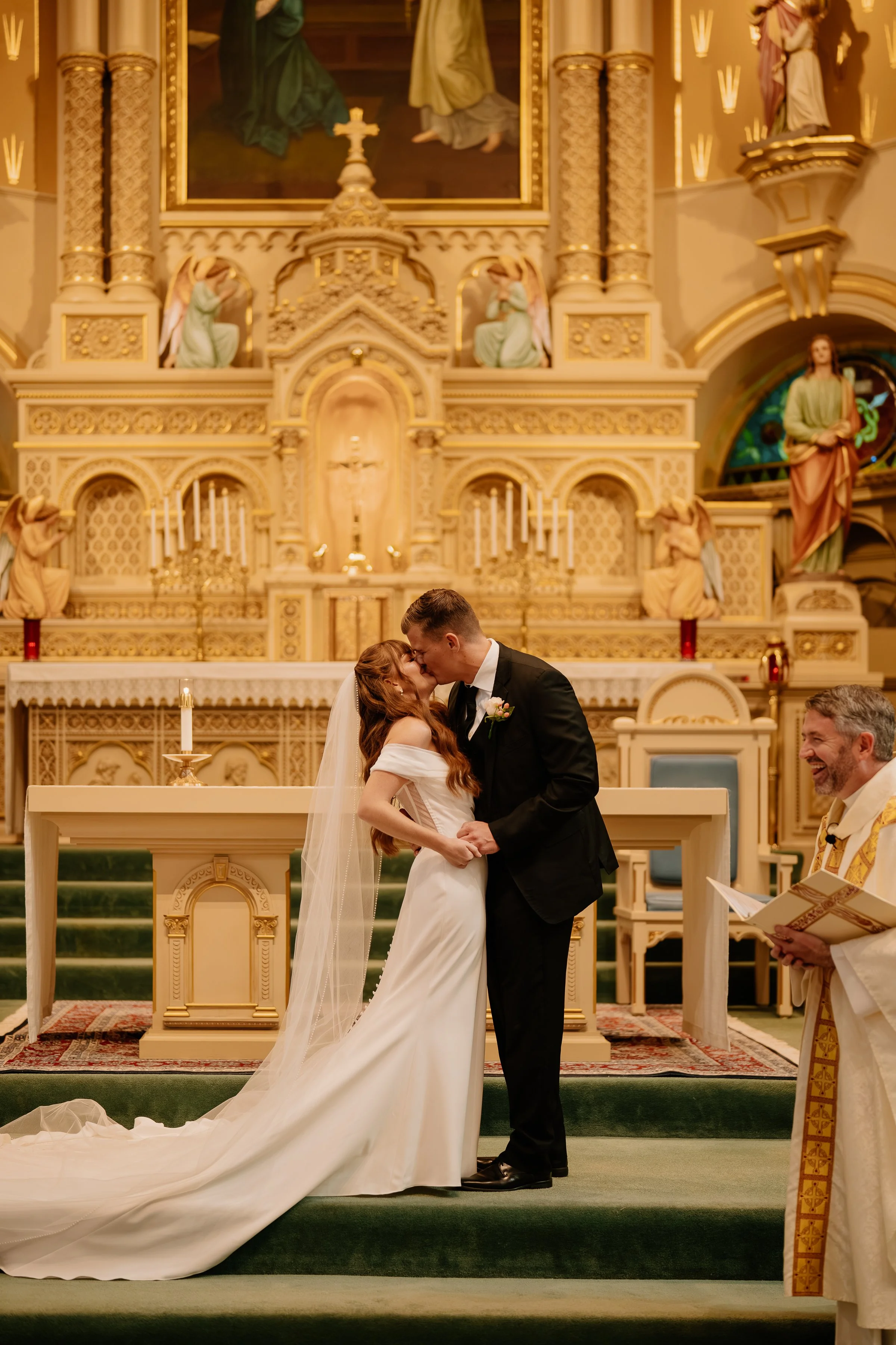 A bride and groom sharing a kiss at the altar during a wedding ceremony in a church with ornate gold decorations and religious statues.