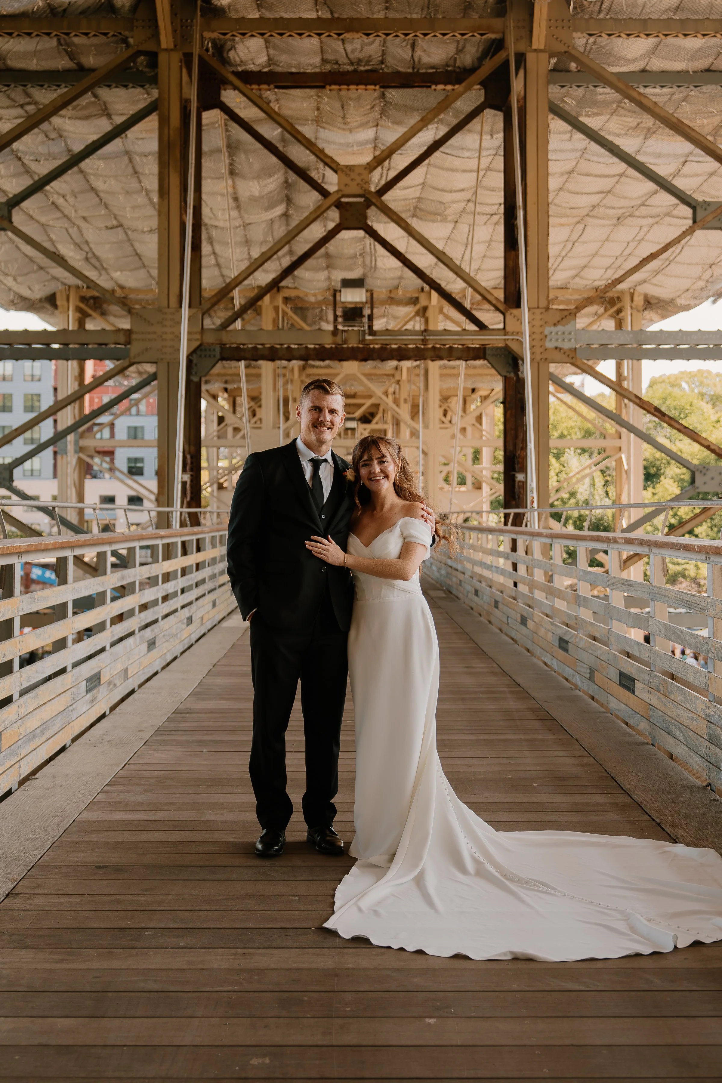 A bride and groom standing together on a wooden walkway, smiling. The bride is in a white wedding gown with a train, and the groom is in a black suit. They are outdoors in an industrial-style structure with metal beams and a partially covered ceiling