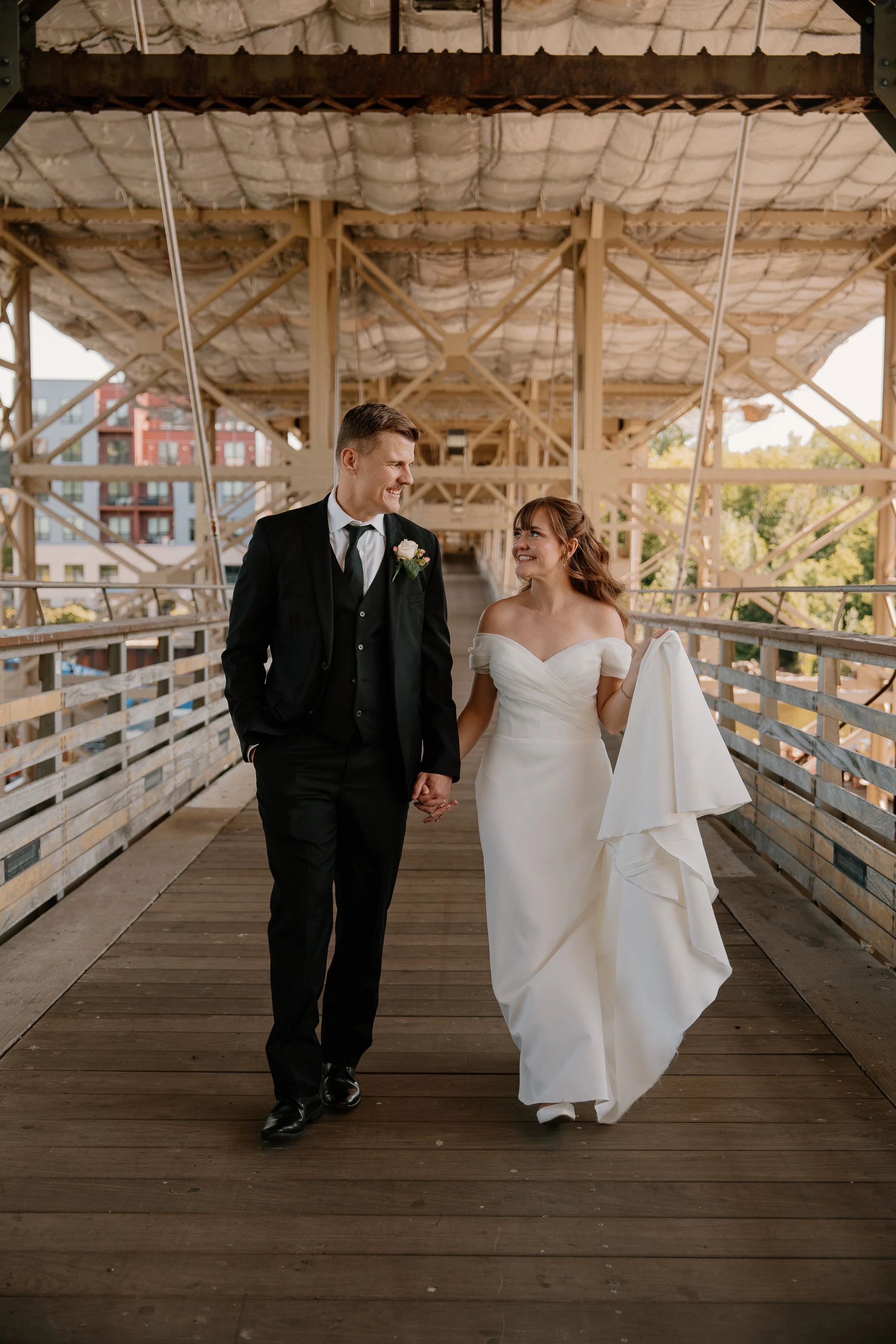 A newly married couple is walking hand in hand on a wooden bridge, smiling at each other, with the bride holding her dress.