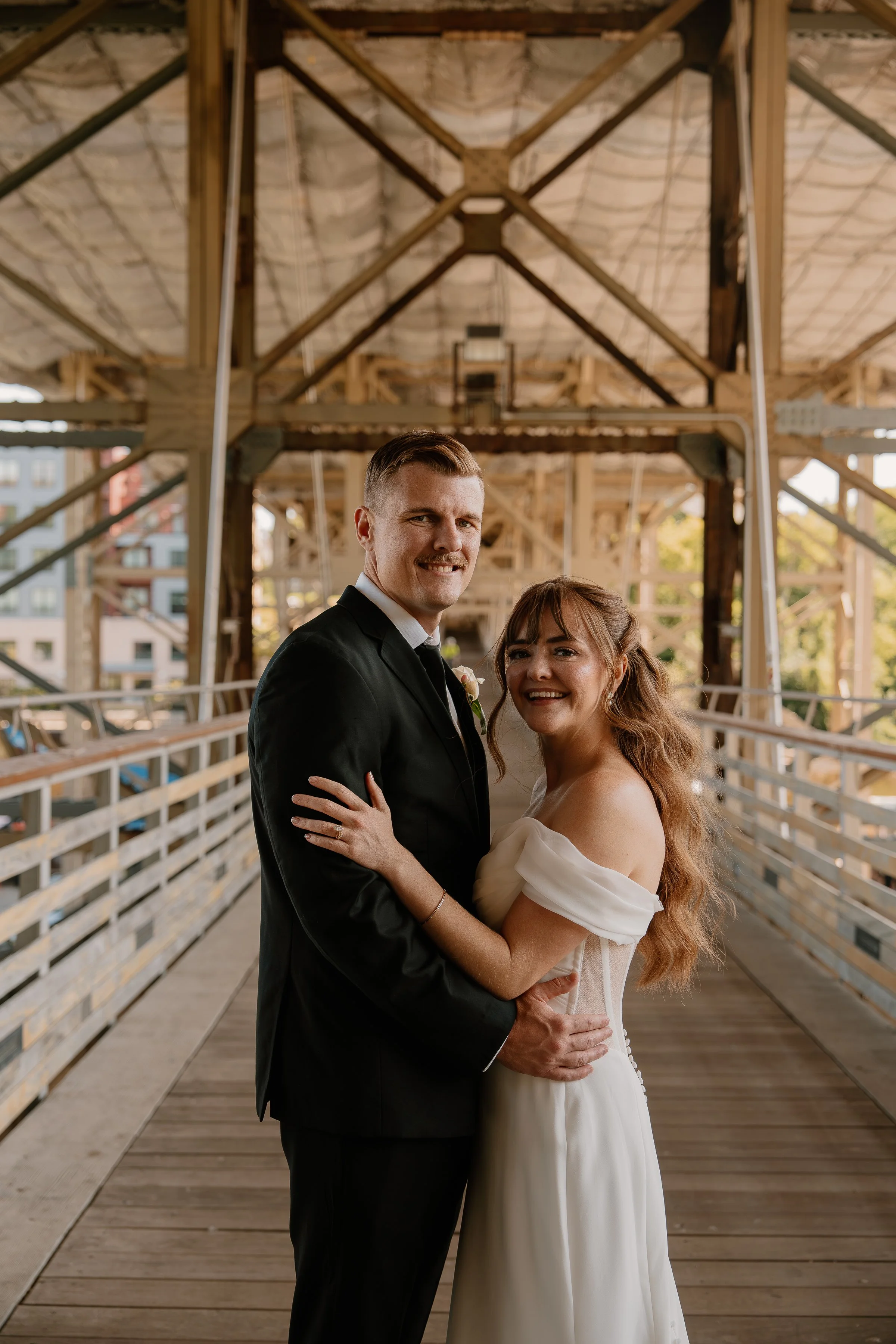 A bride and groom standing together on a wooden bridge, smiling and embracing, with the bride's long red hair and off-the-shoulder wedding dress, and the groom in a black suit and white shirt.