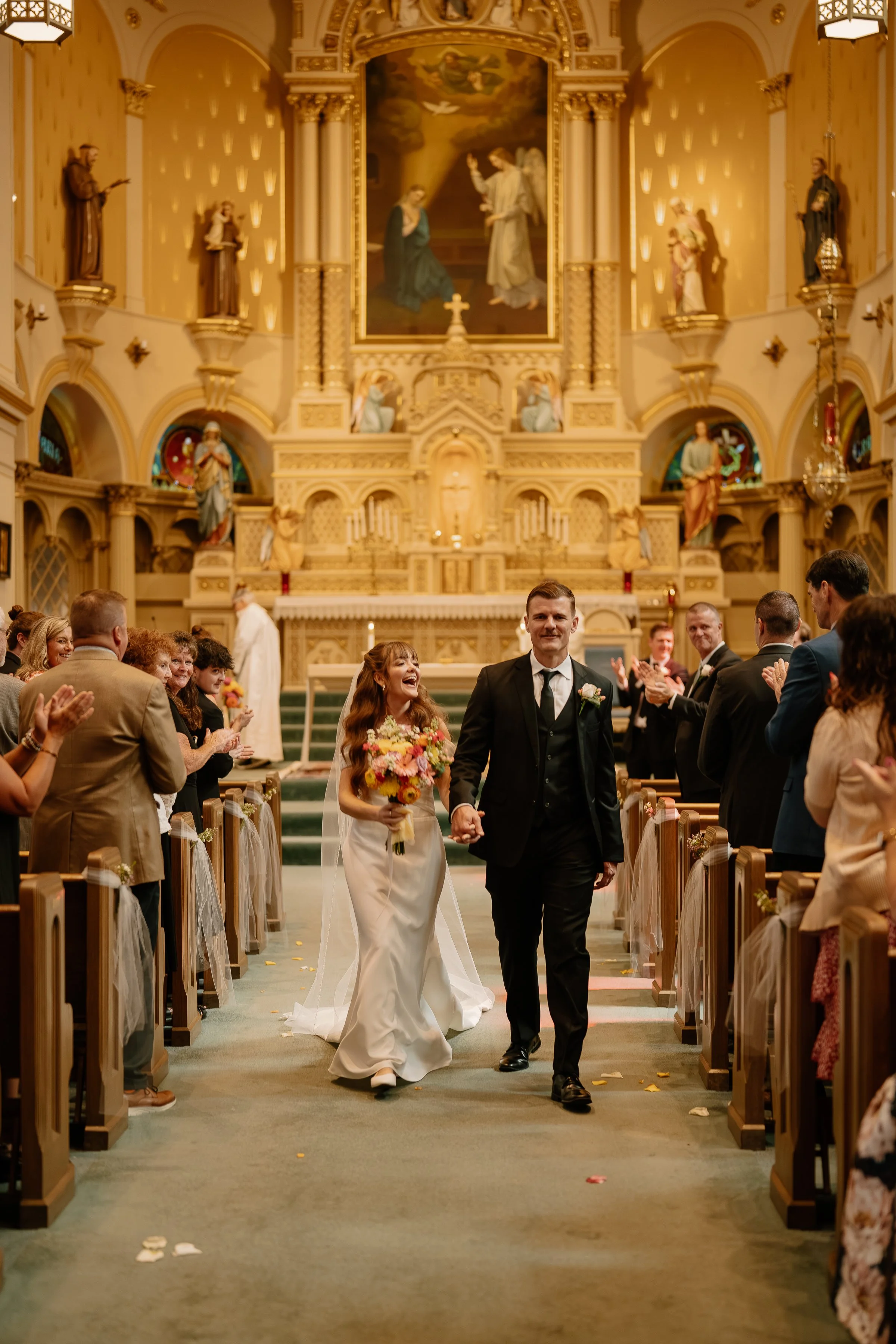 A bride and groom walking down the aisle in a church, surrounded by smiling guests clapping and celebrating after a wedding ceremony.