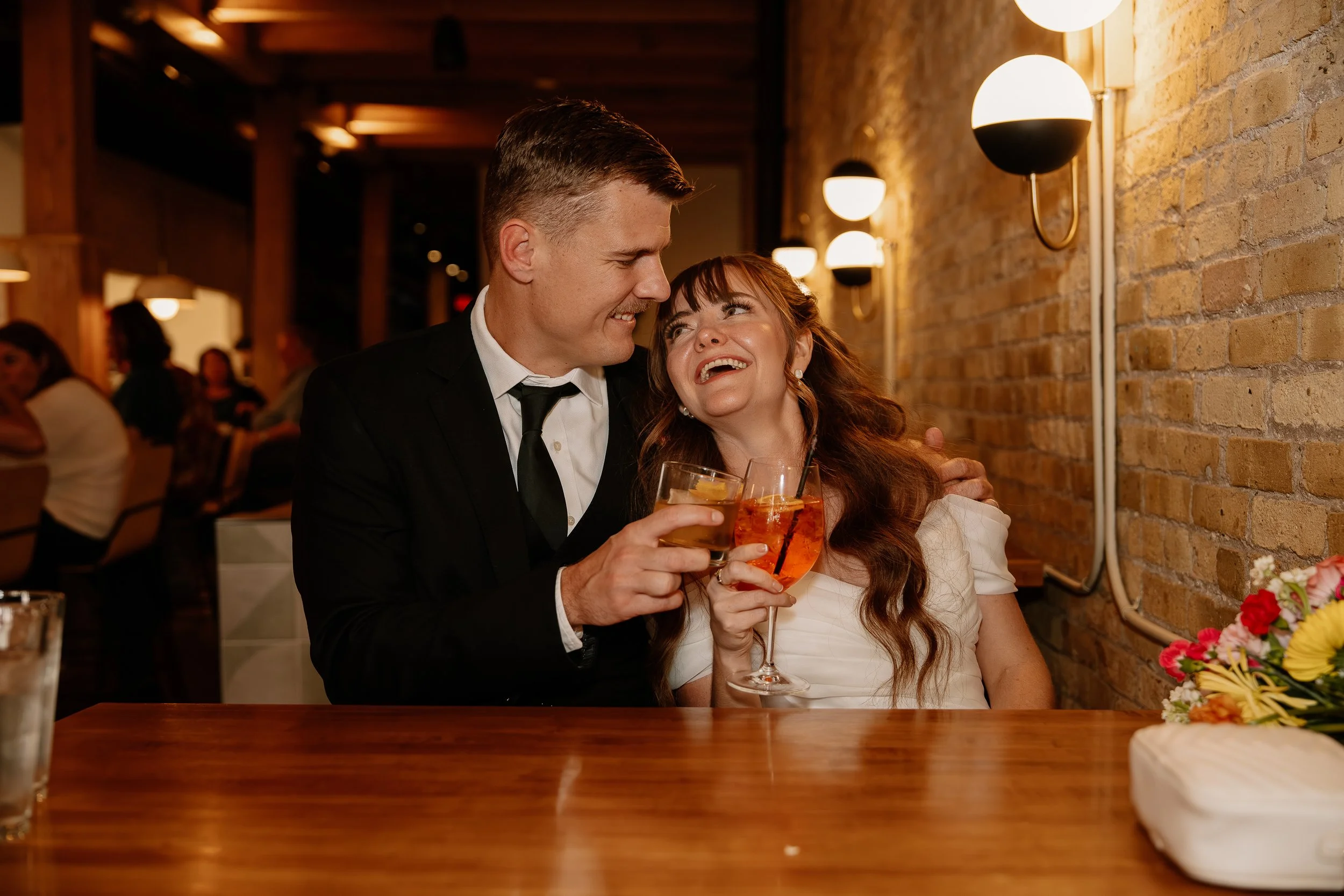 A couple celebrating together at a restaurant, smiling and holding drinks, with a brick wall and warm lighting in the background.