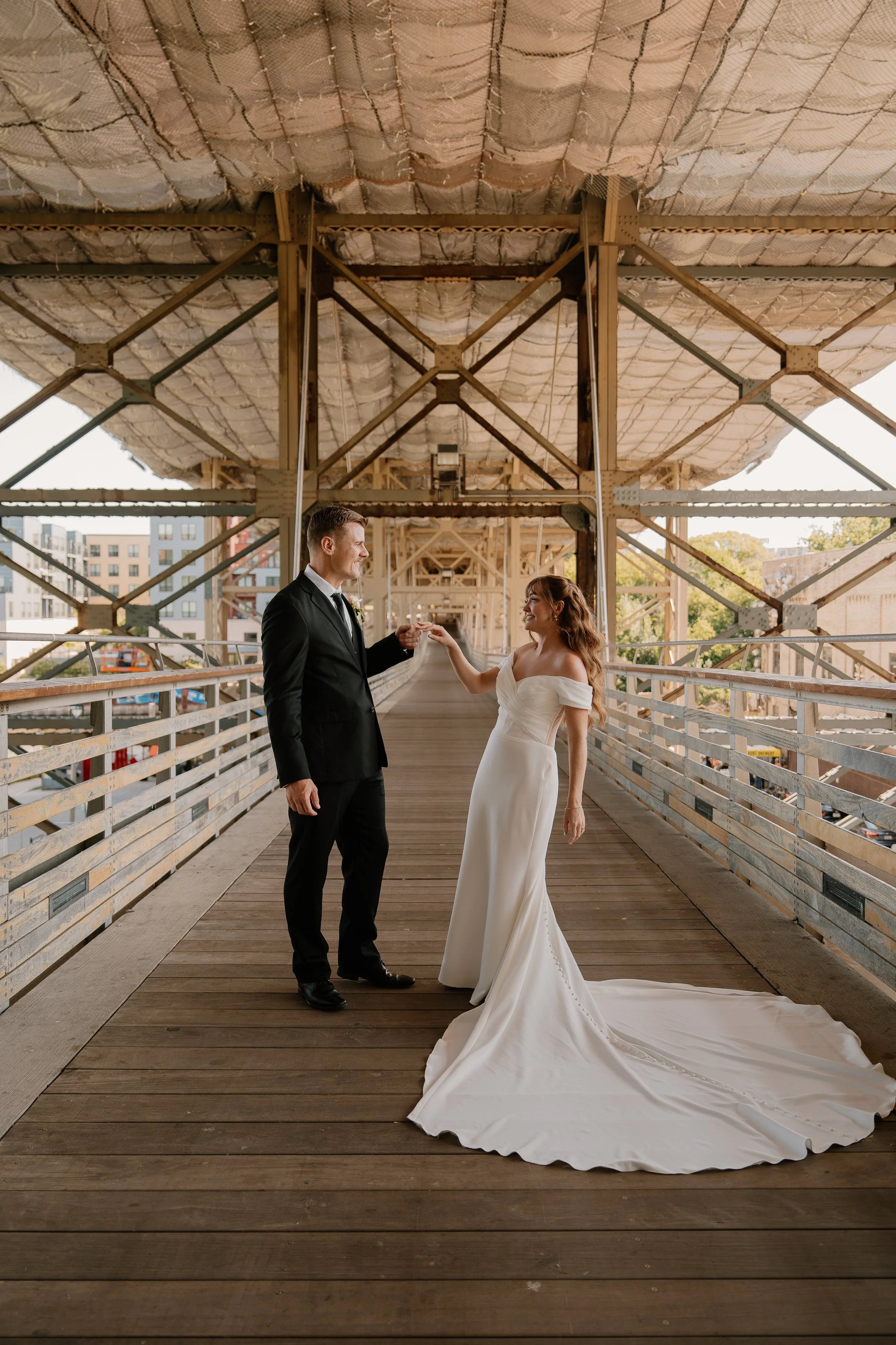 A bride and groom standing on a wooden bridge, holding hands, with the bride in a white wedding gown and the groom in a black suit, under an industrial bridge structure.
