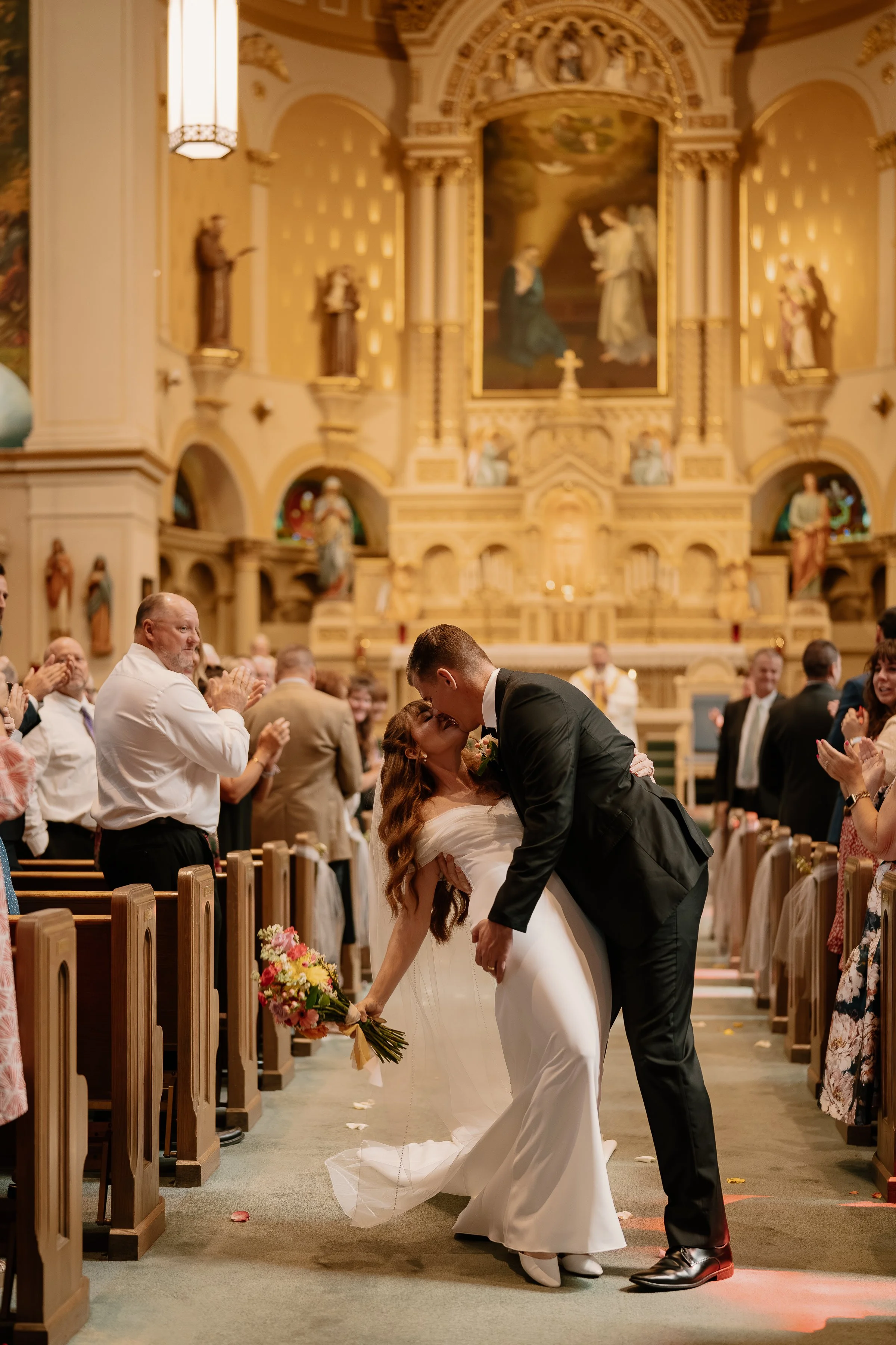 A newlywed couple shares a kiss in a church during their wedding ceremony. The bride holds a bouquet of flowers, and the groom is dipping her. The church is decorated with religious statues and ornate artwork in the background, and guests are clappin