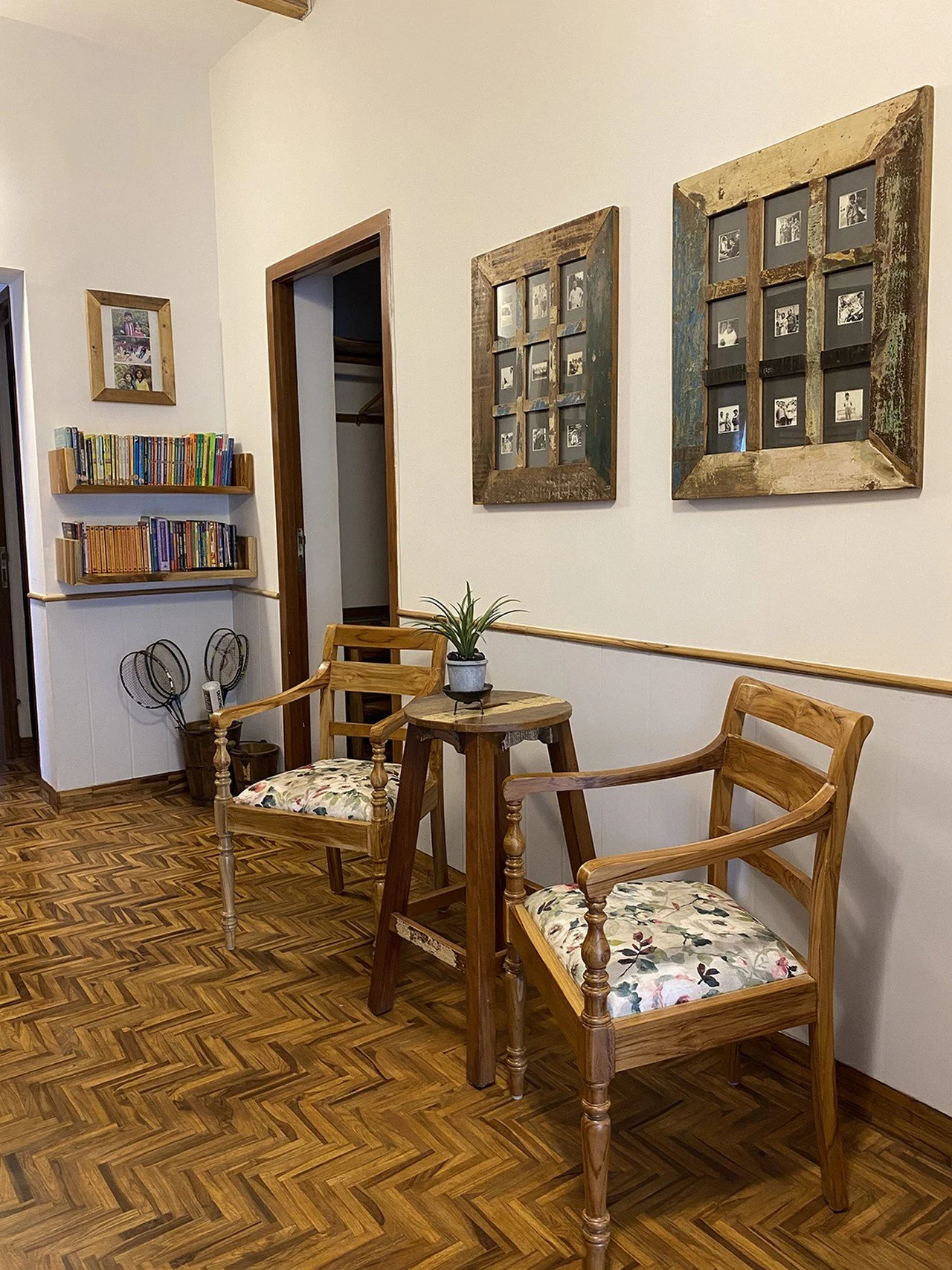 A cozy corner with two wooden chairs with floral cushions, a small round wooden table with a potted plant, a bookshelf filled with colorful books, and framed black and white photographs on the wall. The floor has a herringbone wood pattern.